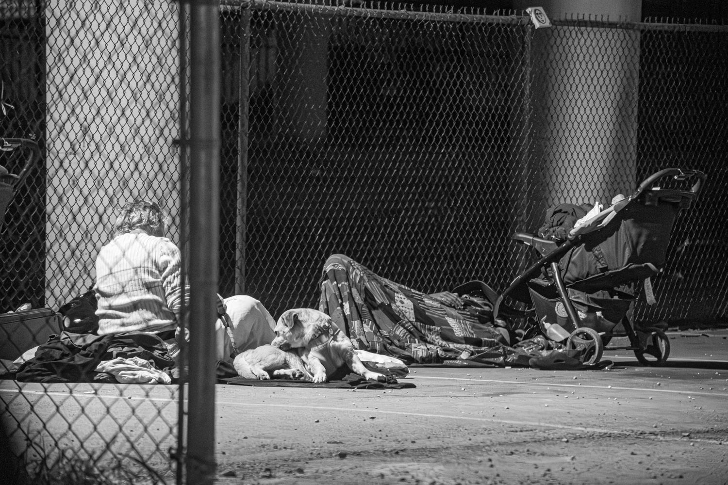 Black and white photo of homeless people sleeping on a sidewalk beside a chain-link fence.
