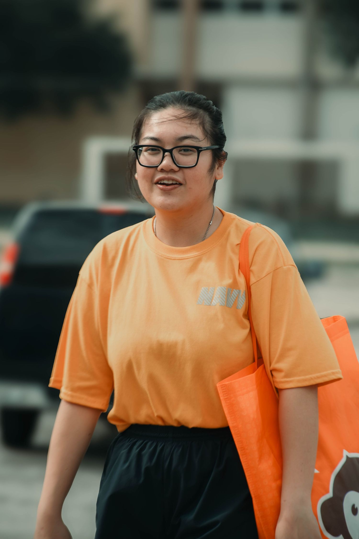 Smiling Asian woman in orange t-shirt wearing glasses carrying orange bag outdoors.