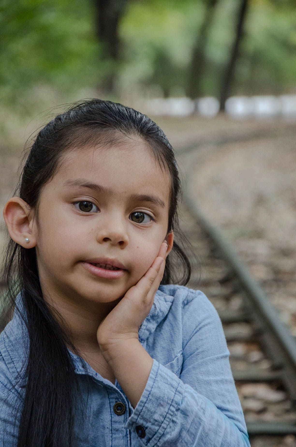 Young girl with dark hair rests her cheek on her hand near railroad tracks in a park setting.