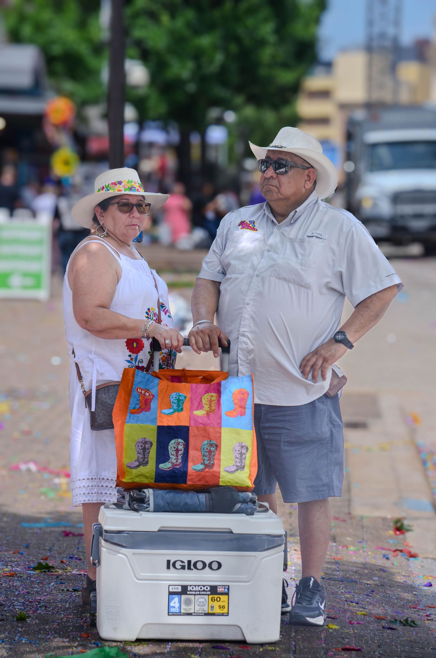 Couple standing next to Igloo cooler at outdoor festival wearing hats and sunglasses.
