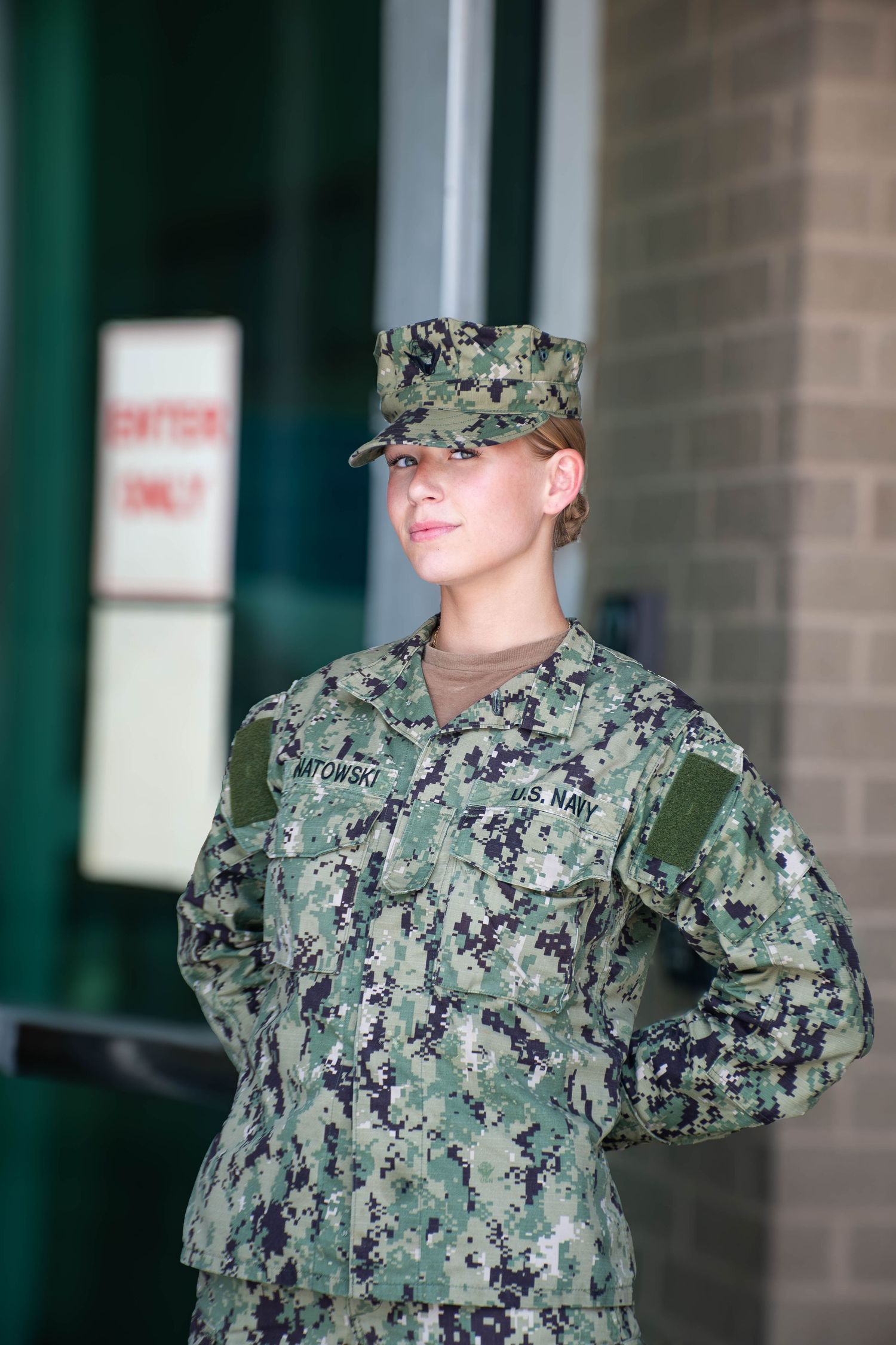 Female US Navy sailor in digital camouflage uniform and cap standing at attention outdoors.