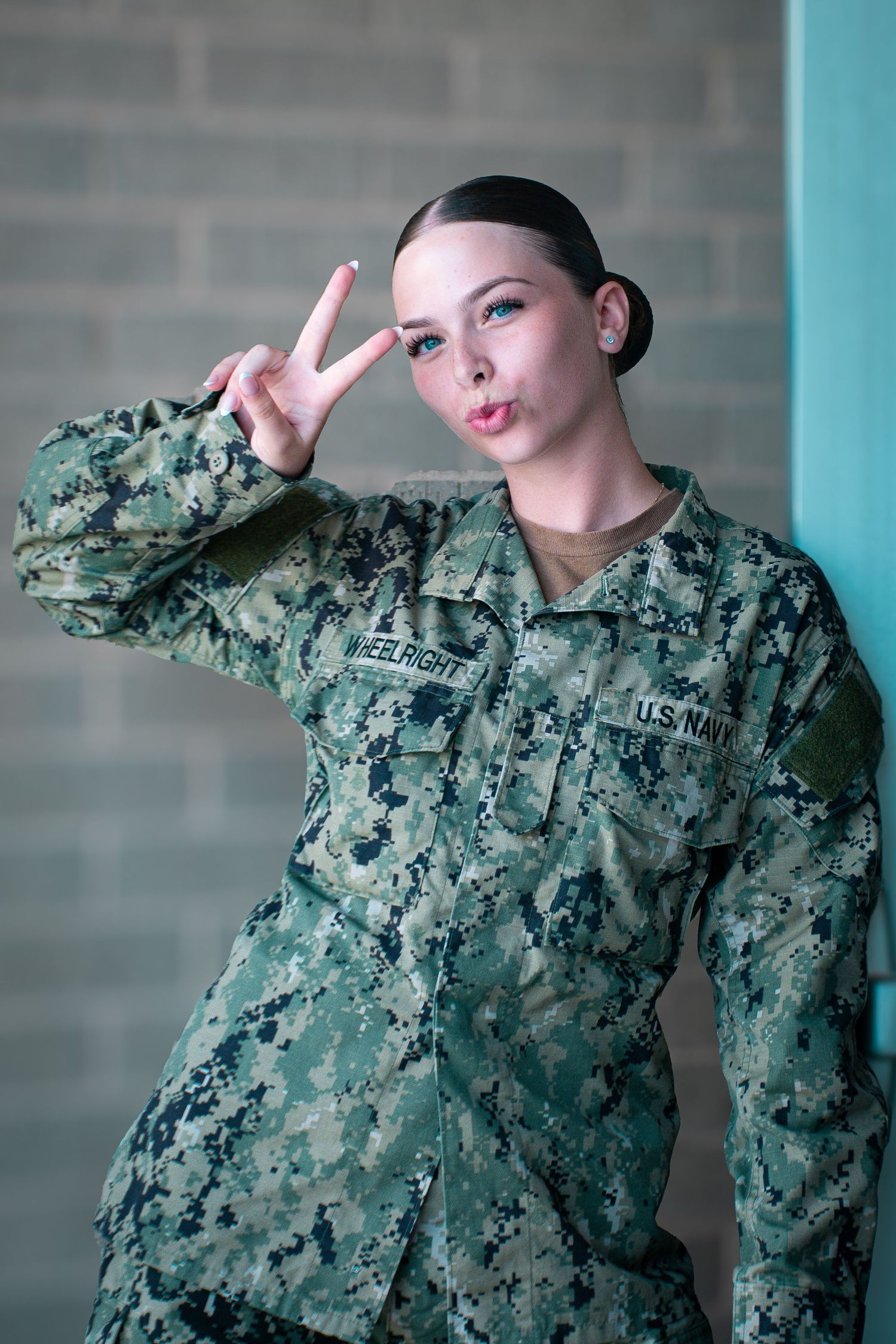 Female US Navy sailor in camouflage uniform making a peace sign gesture against a teal wall.