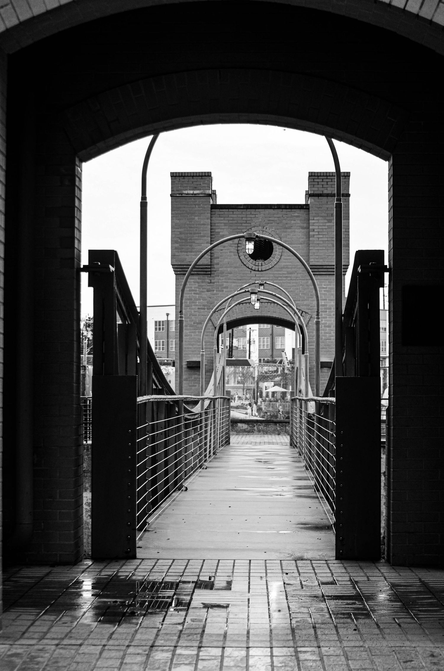 Black and white view through arched brick gateway leading to a pedestrian suspension bridge in the city.