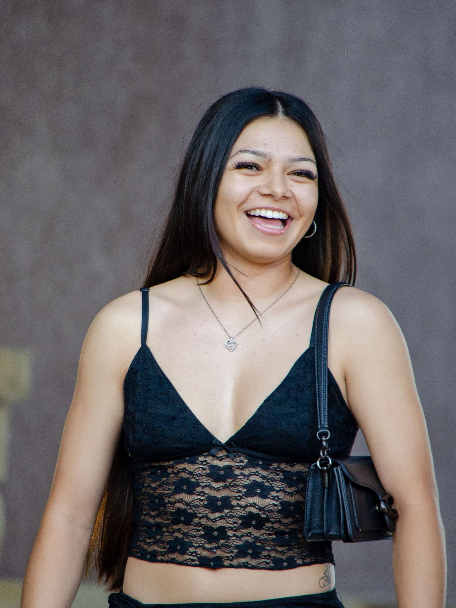 Laughing young woman with long black hair in black lace crop top holding a small purse.