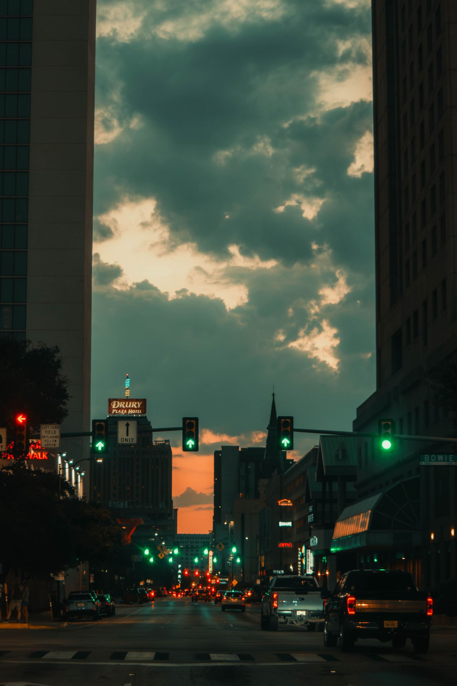 Downtown city street at dusk with green traffic lights and dramatic cloudy sunset sky.