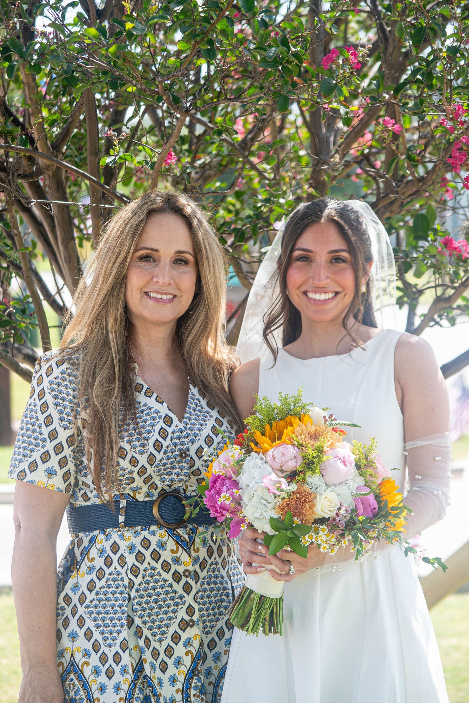 Bride in white dress with veil holding colorful bouquet posing with woman outdoors.