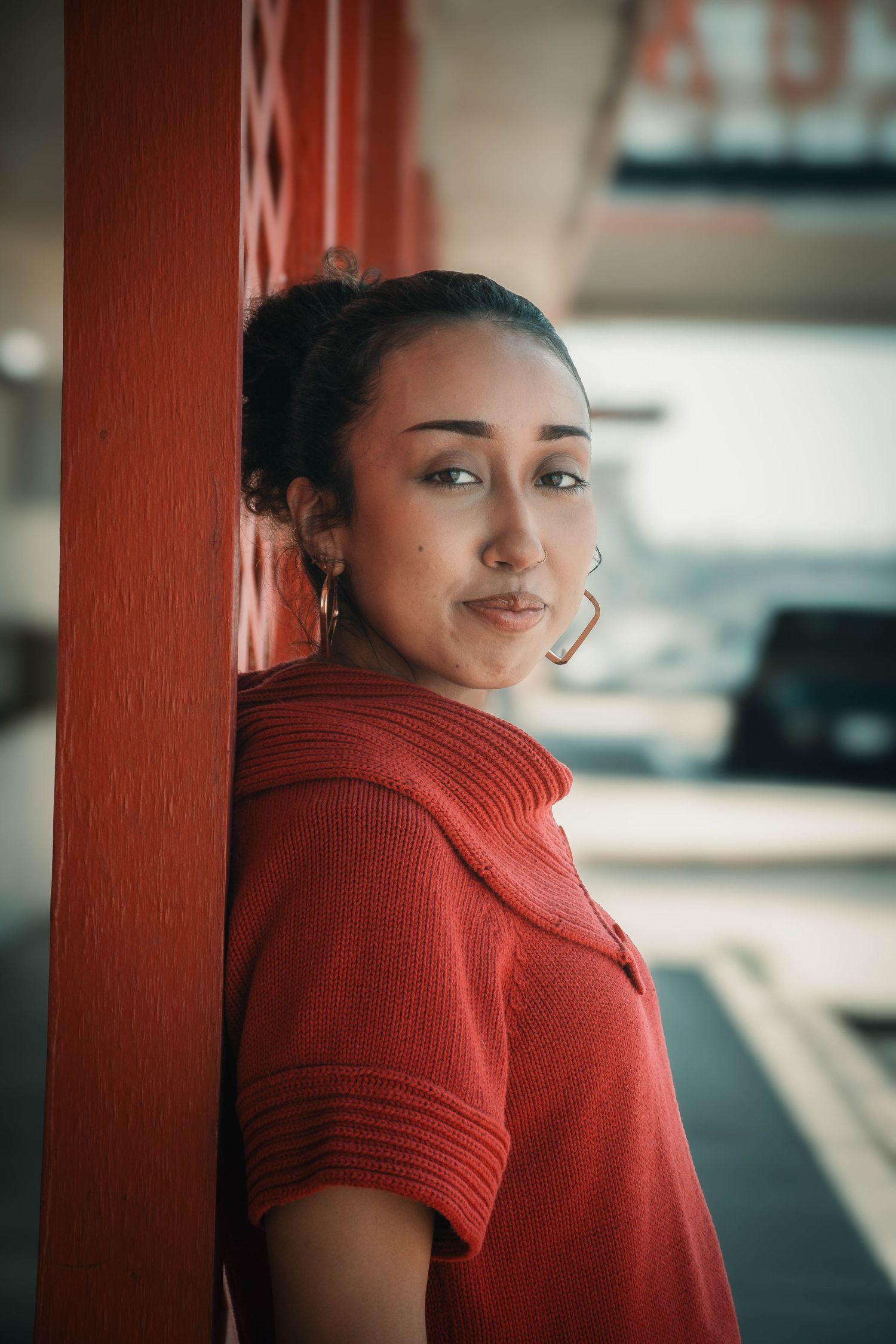 Young woman in red sweater leaning against a red pillar, gazing at camera with a subtle smile outdoors.