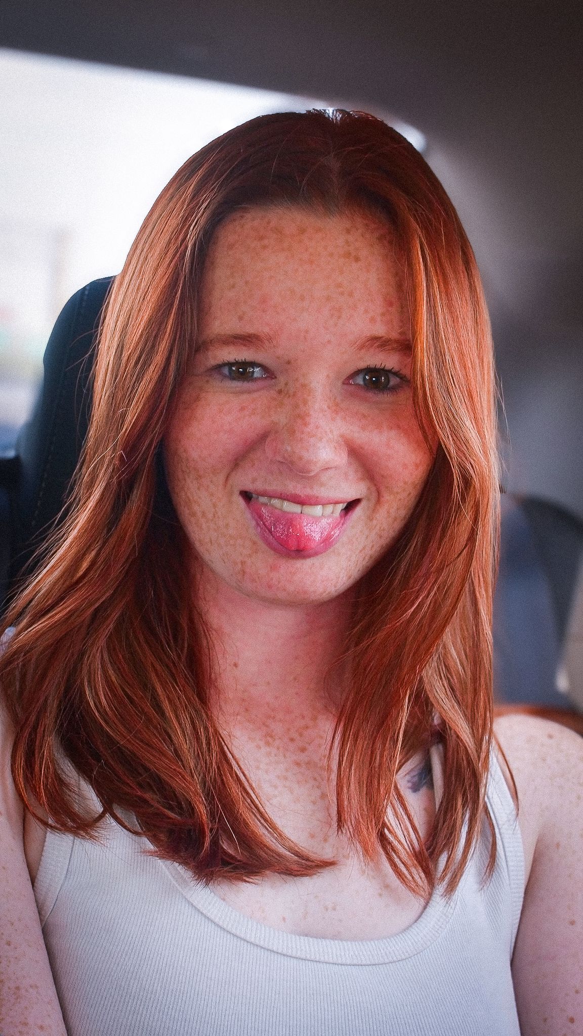 Smiling redhead woman with freckles sticking out tongue, sitting in a car wearing a white tank top.