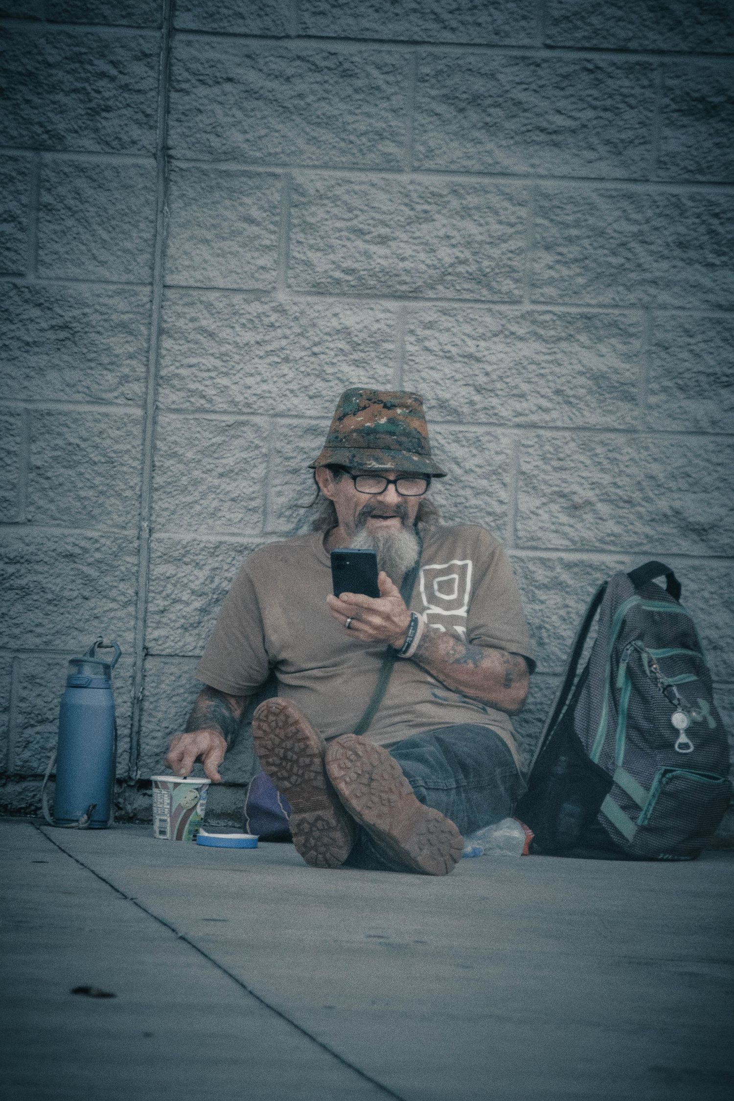 Homeless man sitting against a stone wall on a sidewalk, smiling at his smartphone, with a backpack and cup nearby.