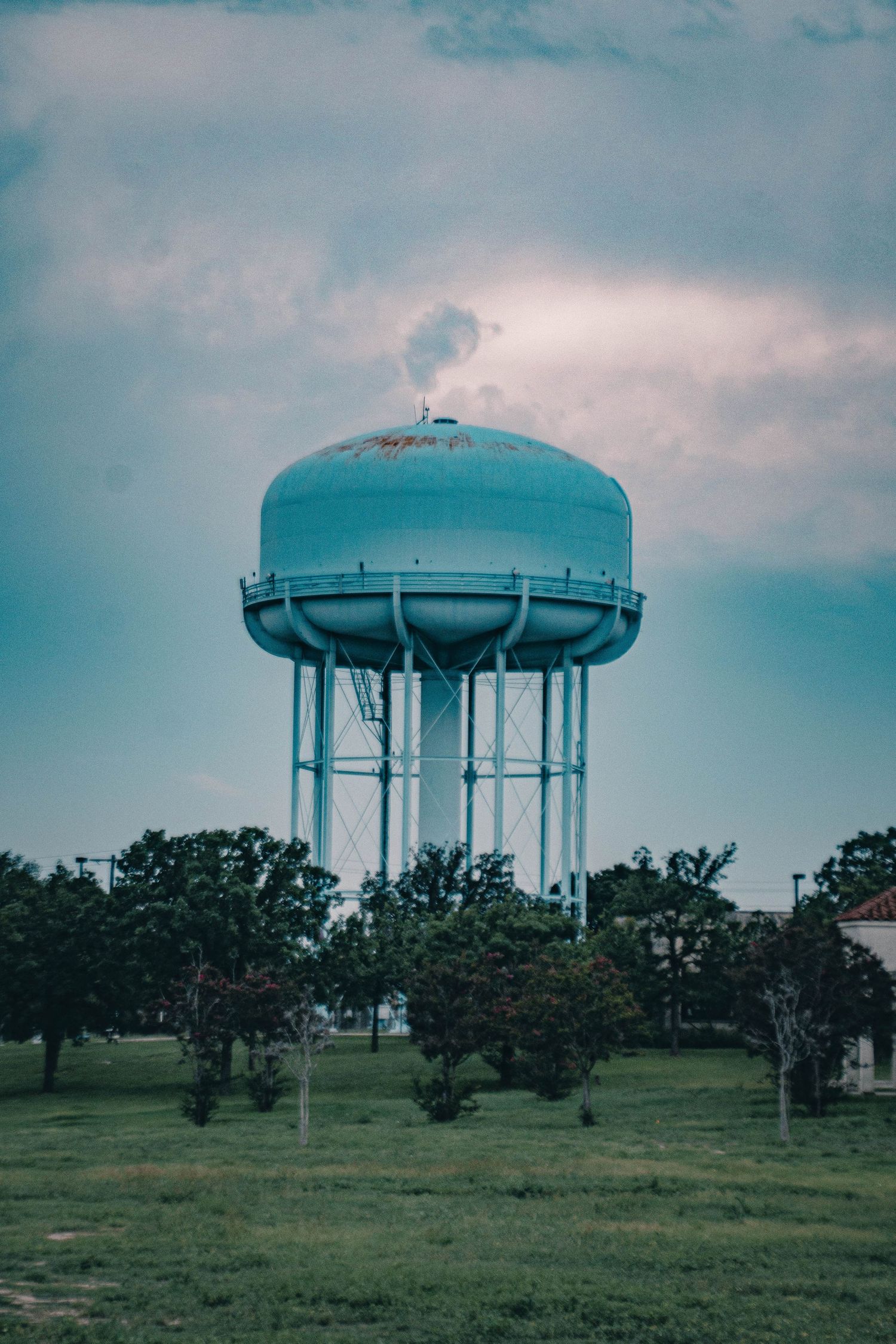 Light blue water tower rising above green trees against a cloudy sky in a grassy field.