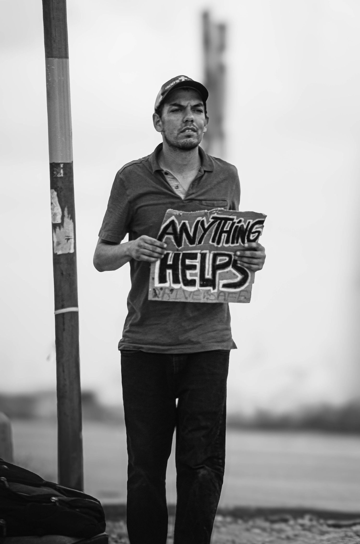 Black and white photo of a man holding an Anything Helps sign while standing on a street corner.
