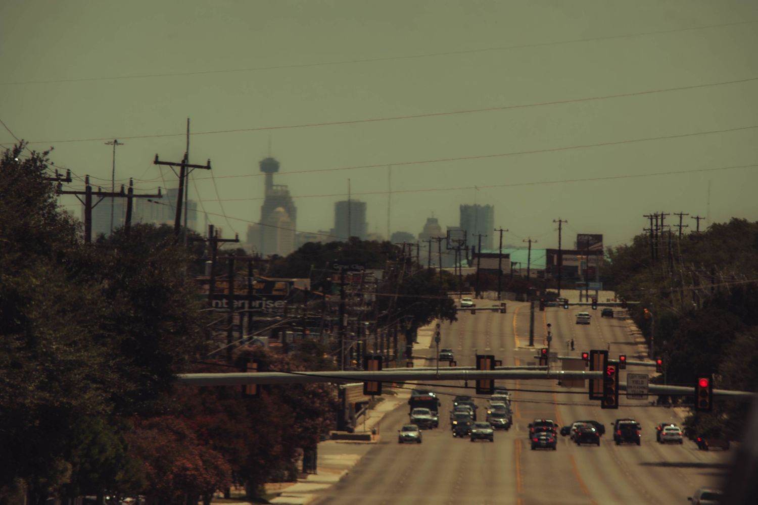 Cars driving on a multi-lane urban highway toward a hazy city skyline with tall buildings and utility poles lining the road.