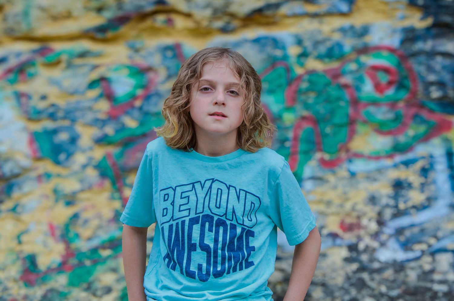 Young blonde boy in teal Beyond Awesome t-shirt standing in front of colorful graffiti wall.