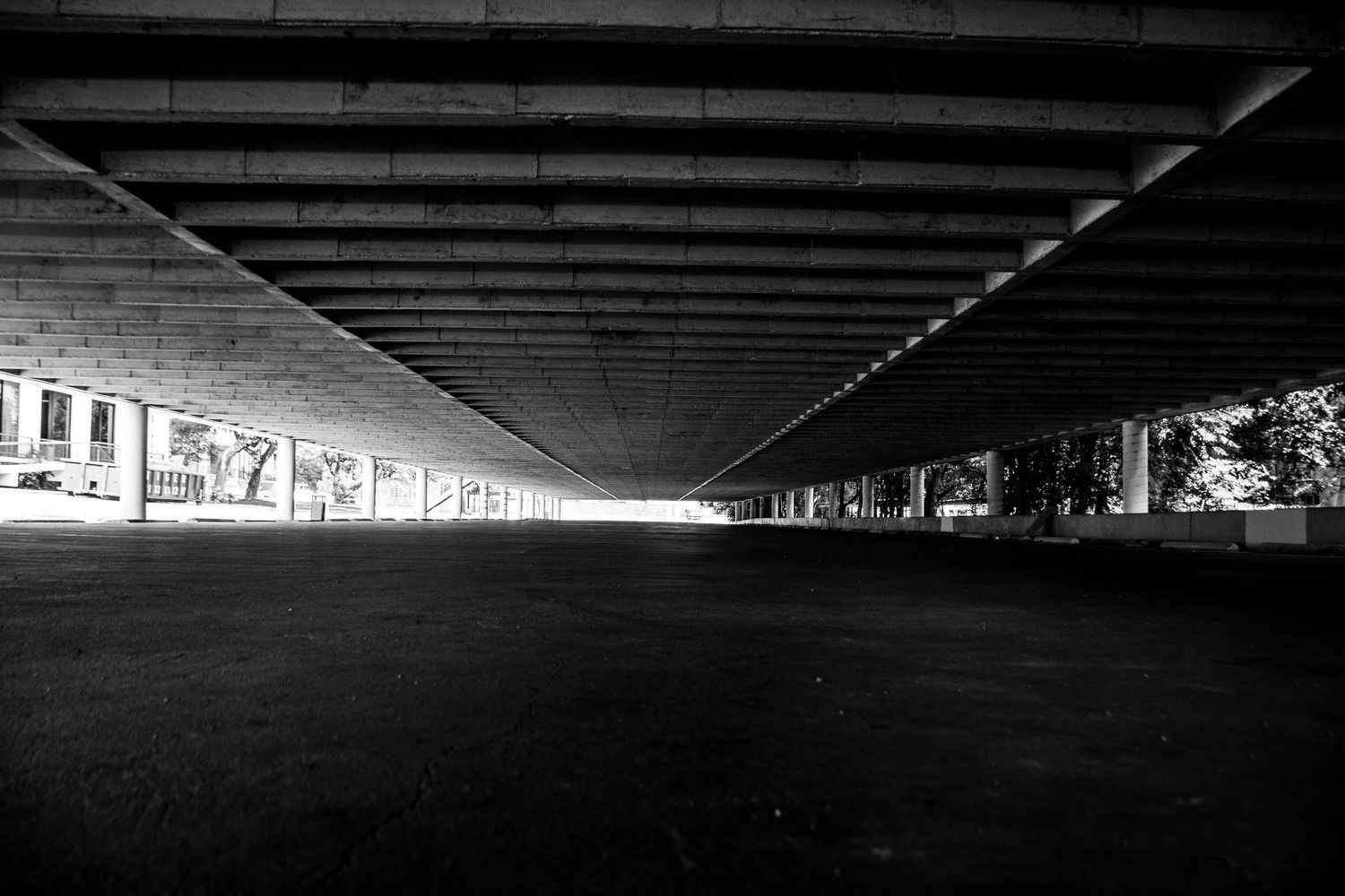 Black and white symmetrical view under a wide concrete bridge overpass with dramatic light visible at the far end.