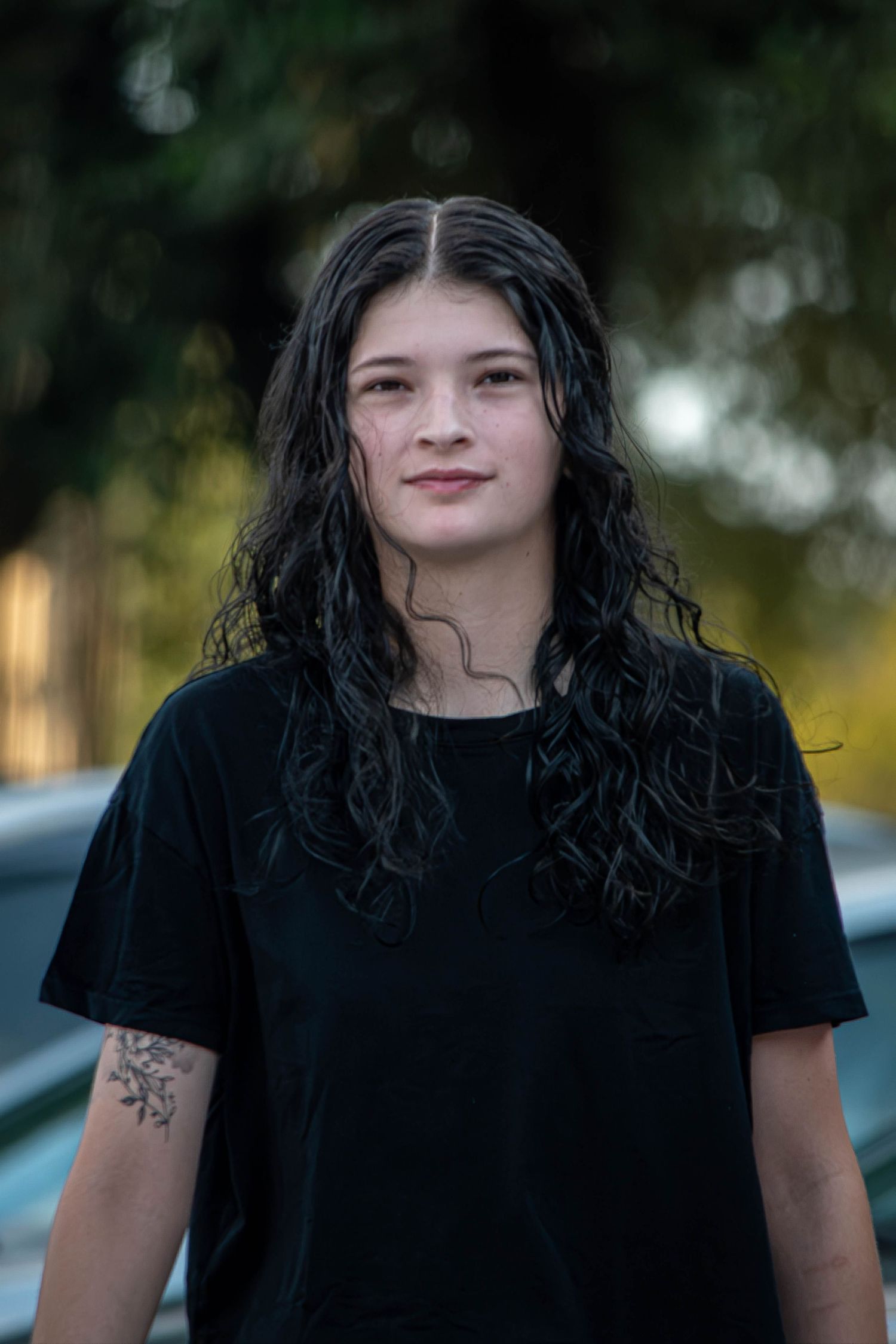 Young woman with long dark curly wet hair wearing a black t-shirt with a floral arm tattoo outdoors.