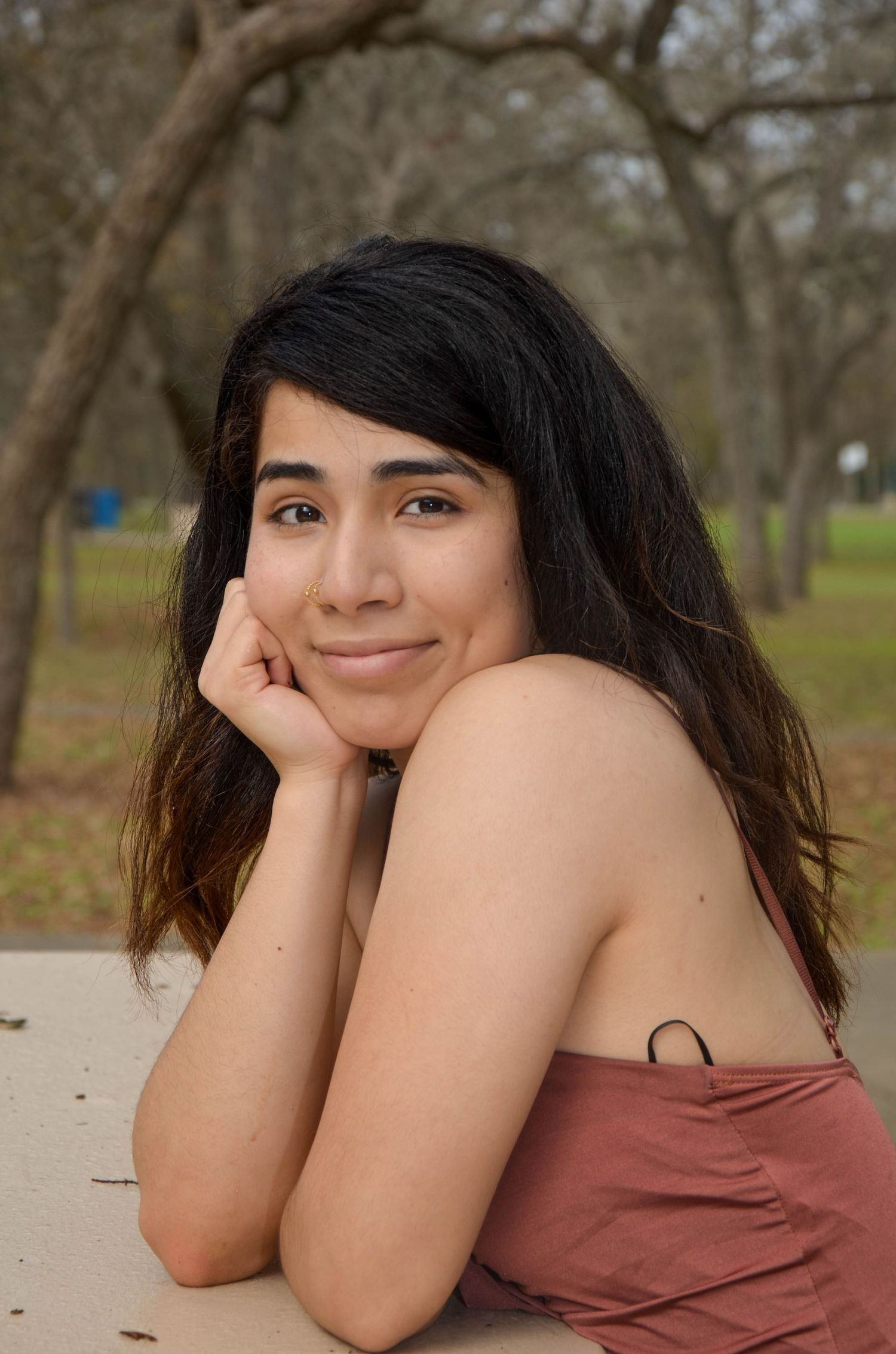 Young woman with dark hair resting chin on hands at outdoor table with bare trees behind her.