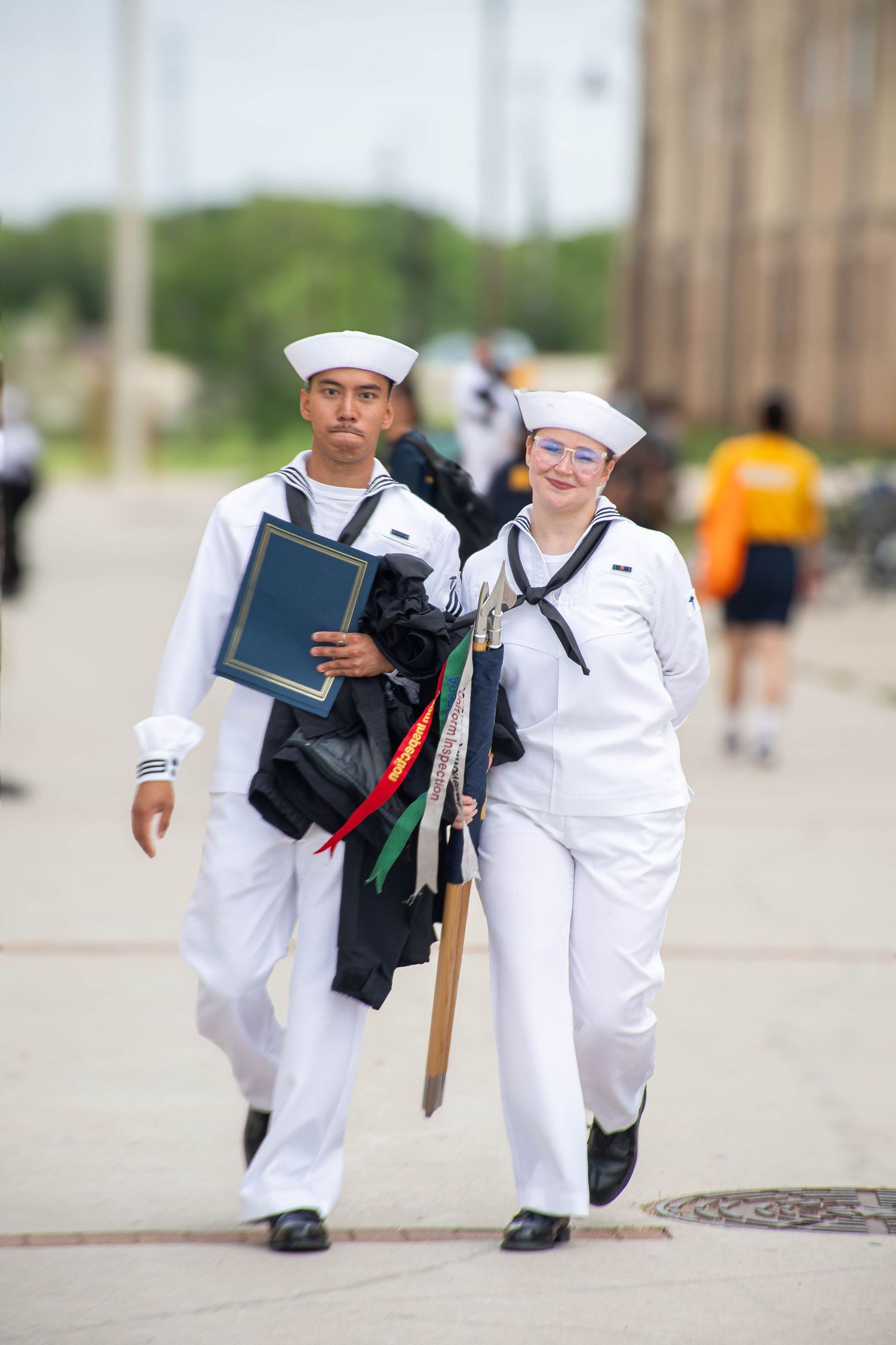Two Navy sailors in white uniforms walk together, one carrying a diploma and gear after a graduation ceremony.