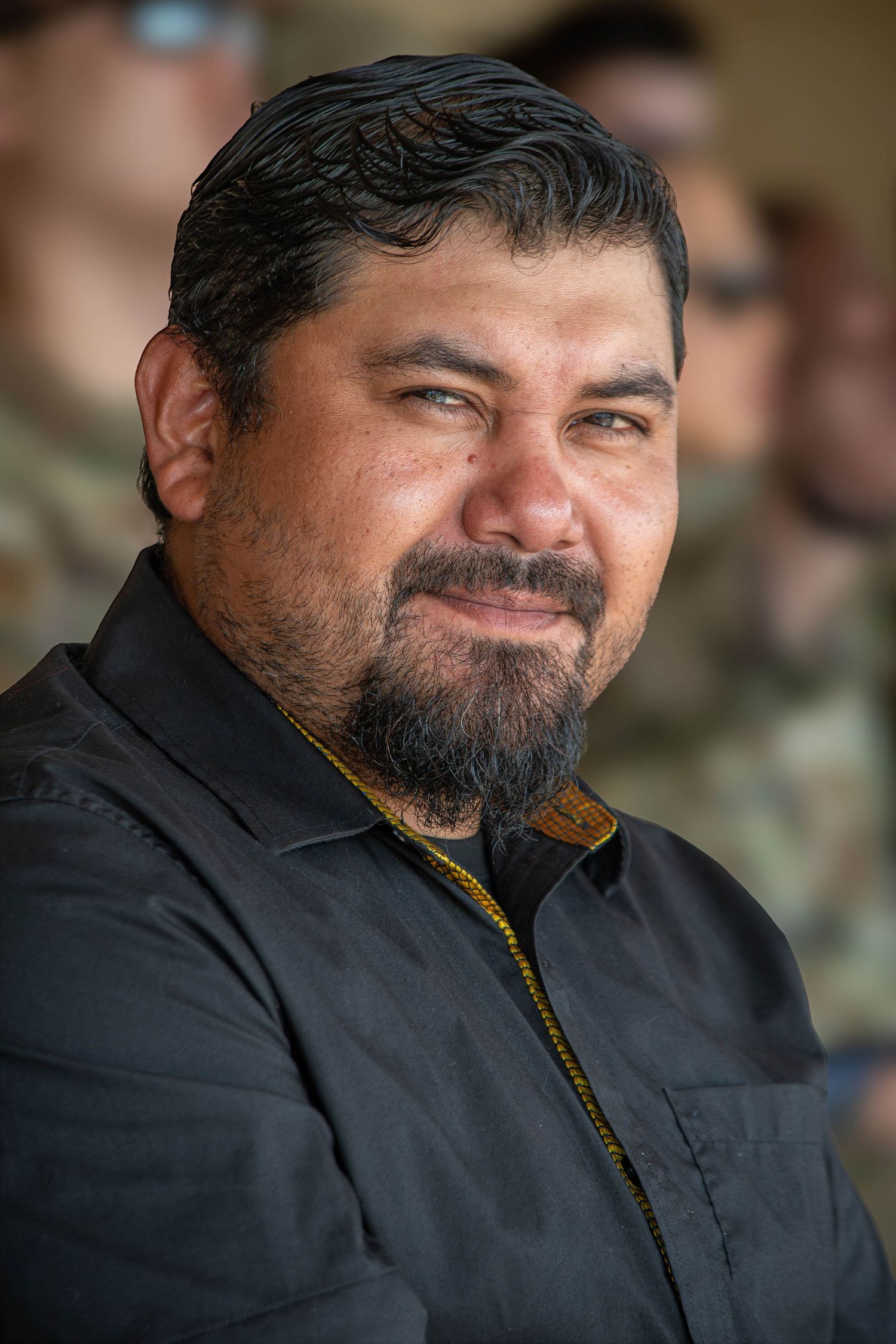 A bearded Hispanic man in a black jacket smiles warmly at an outdoor military event.