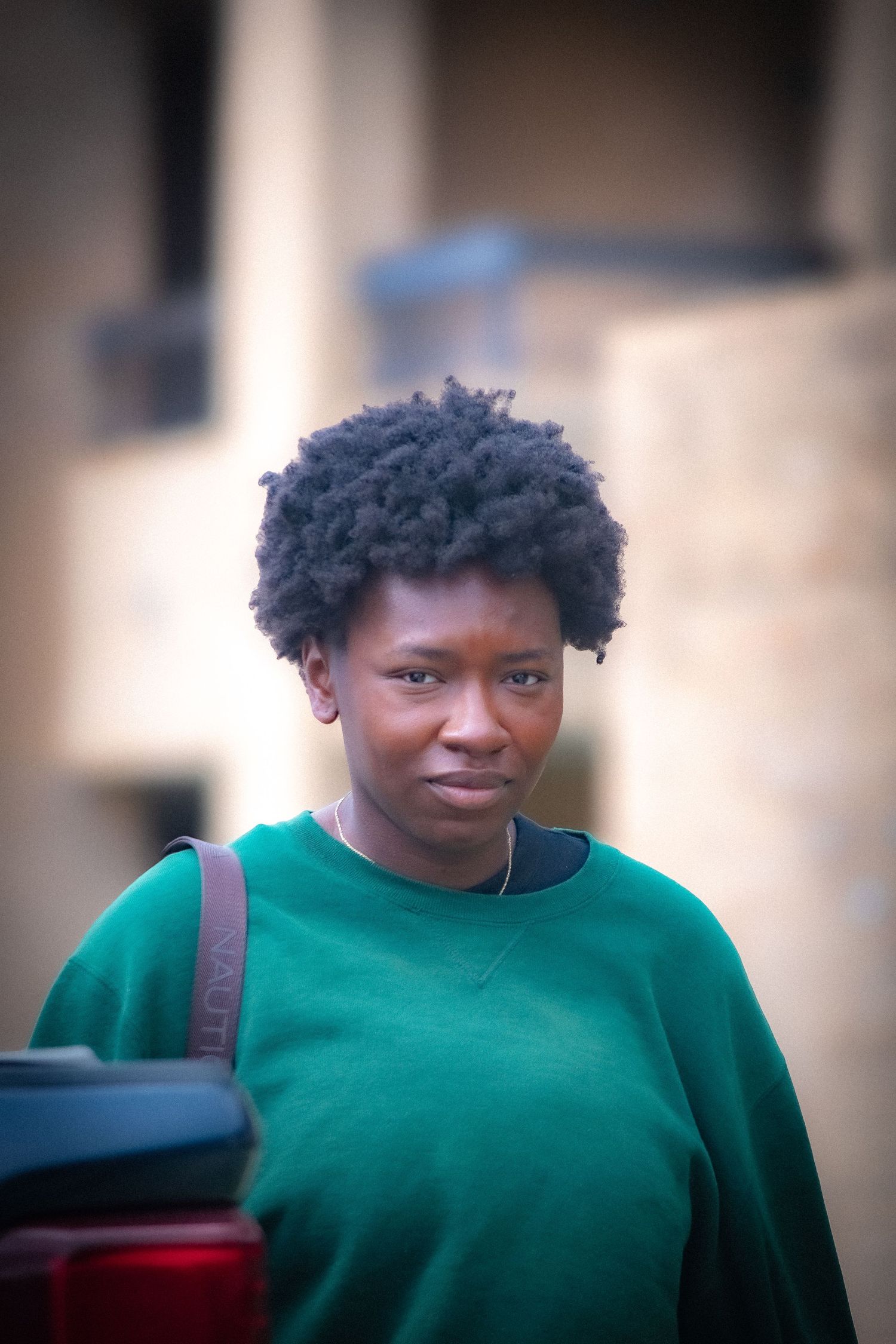 Young Black woman with natural afro hair wearing a green sweatshirt, carrying a bag outdoors.