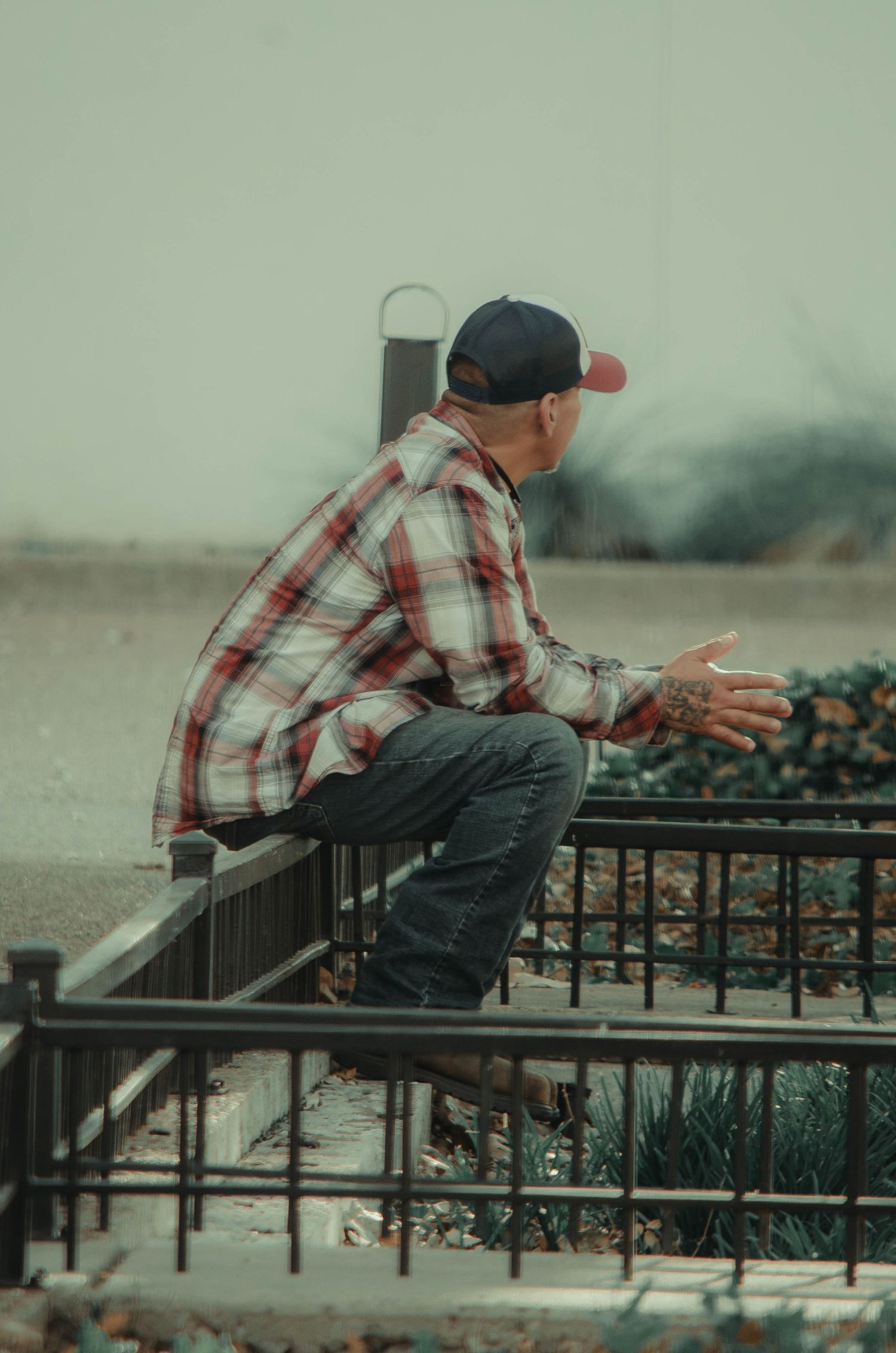 A tattooed man in a plaid shirt and cap sitting on a metal railing outdoors on an overcast day.