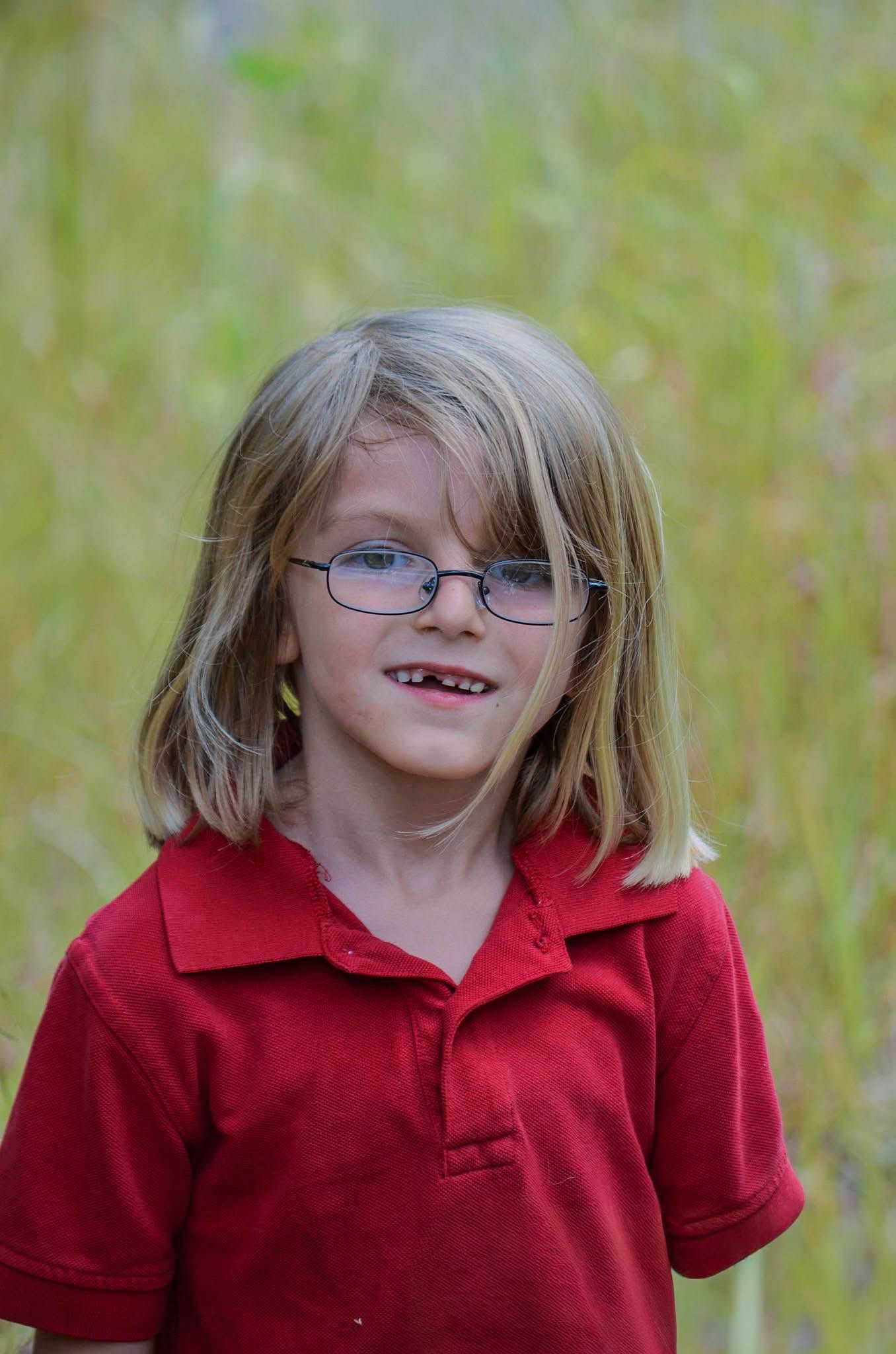 Smiling blonde child with glasses wearing a red polo shirt outdoors.
