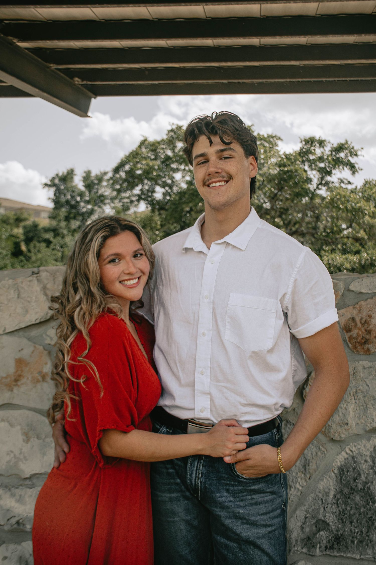 Smiling couple posing together under a covered patio with stone wall and trees behind them.