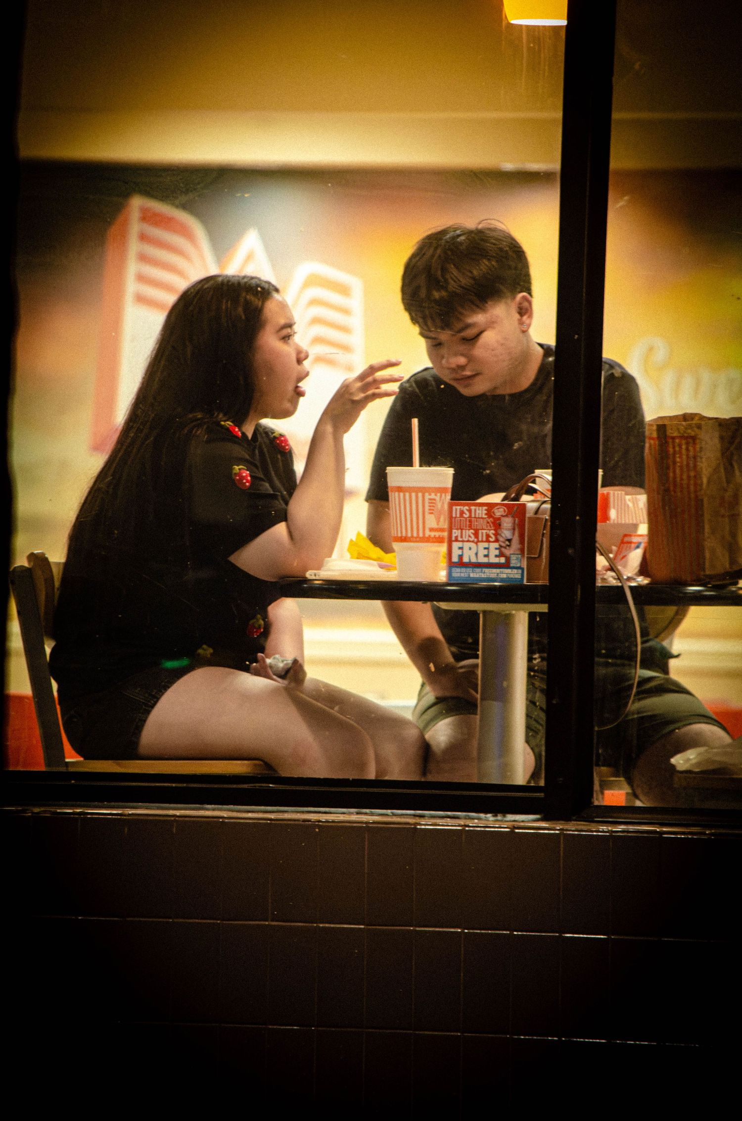 A young couple sharing a conversation over fast food inside a brightly lit restaurant at night.