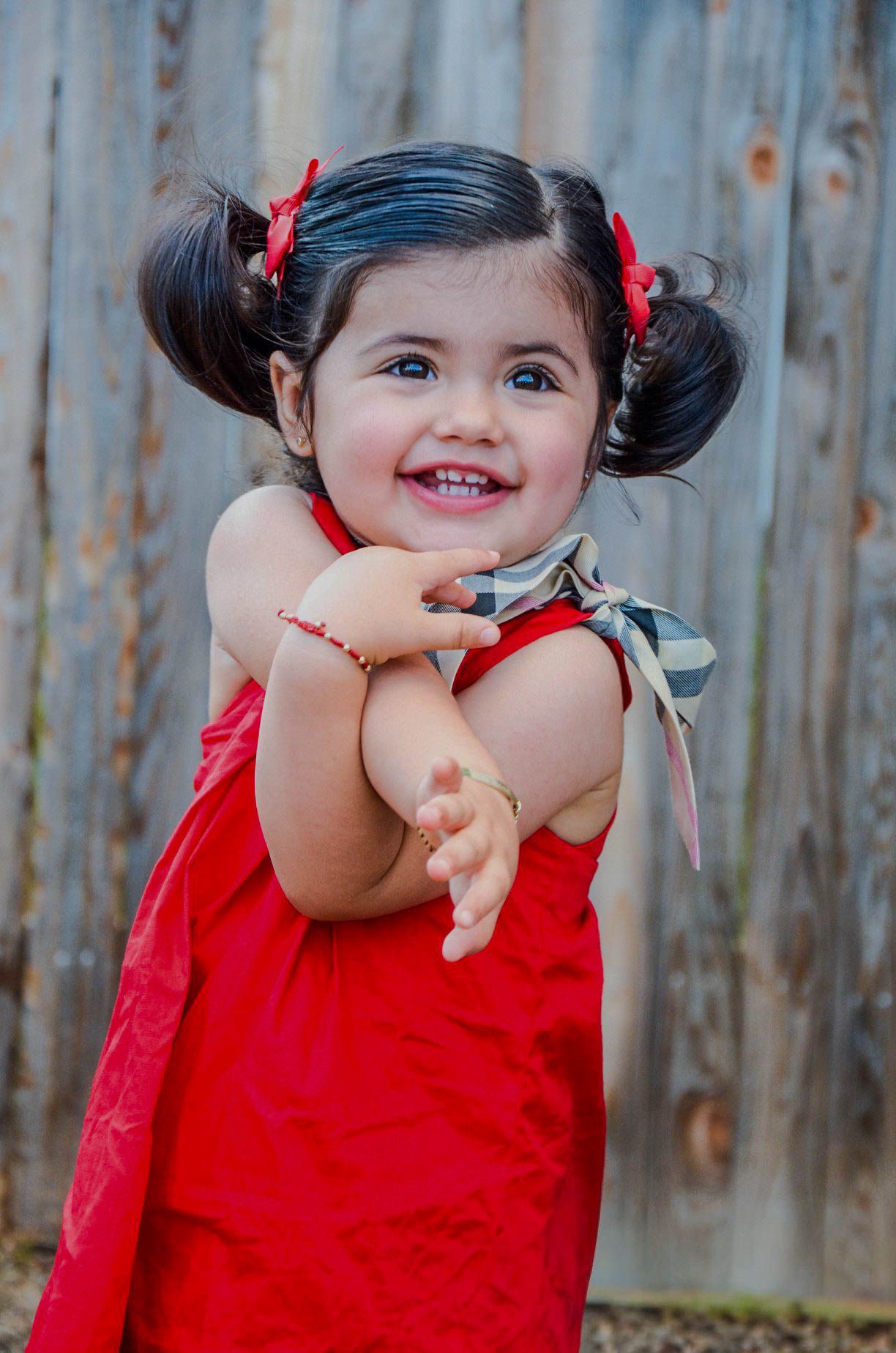 Joyful toddler girl in a red dress with pigtails and red bows smiles brightly outdoors.