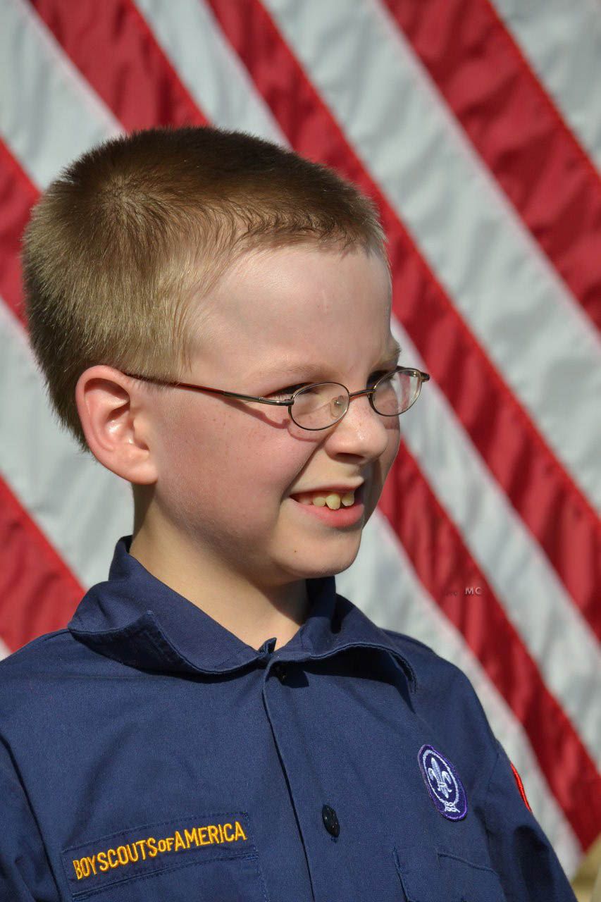 Smiling boy in a blue Boy Scouts of America uniform stands proudly in front of an American flag.