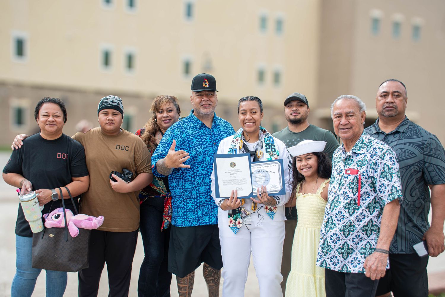 Navy sailor in dress whites celebrates graduation holding certificates with large family group outdoors.