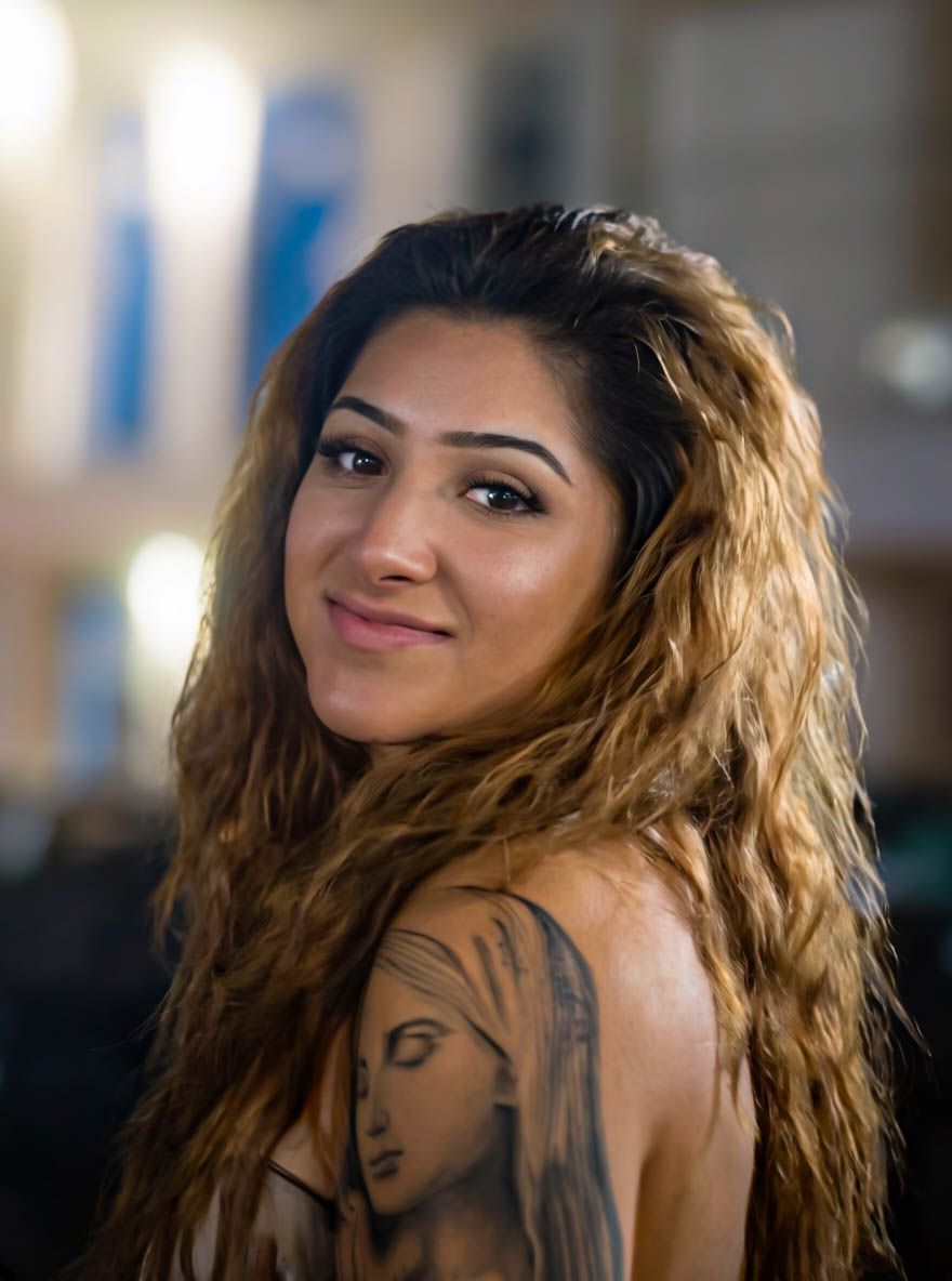 Young woman with long wavy hair and shoulder tattoo smiling over her shoulder indoors.