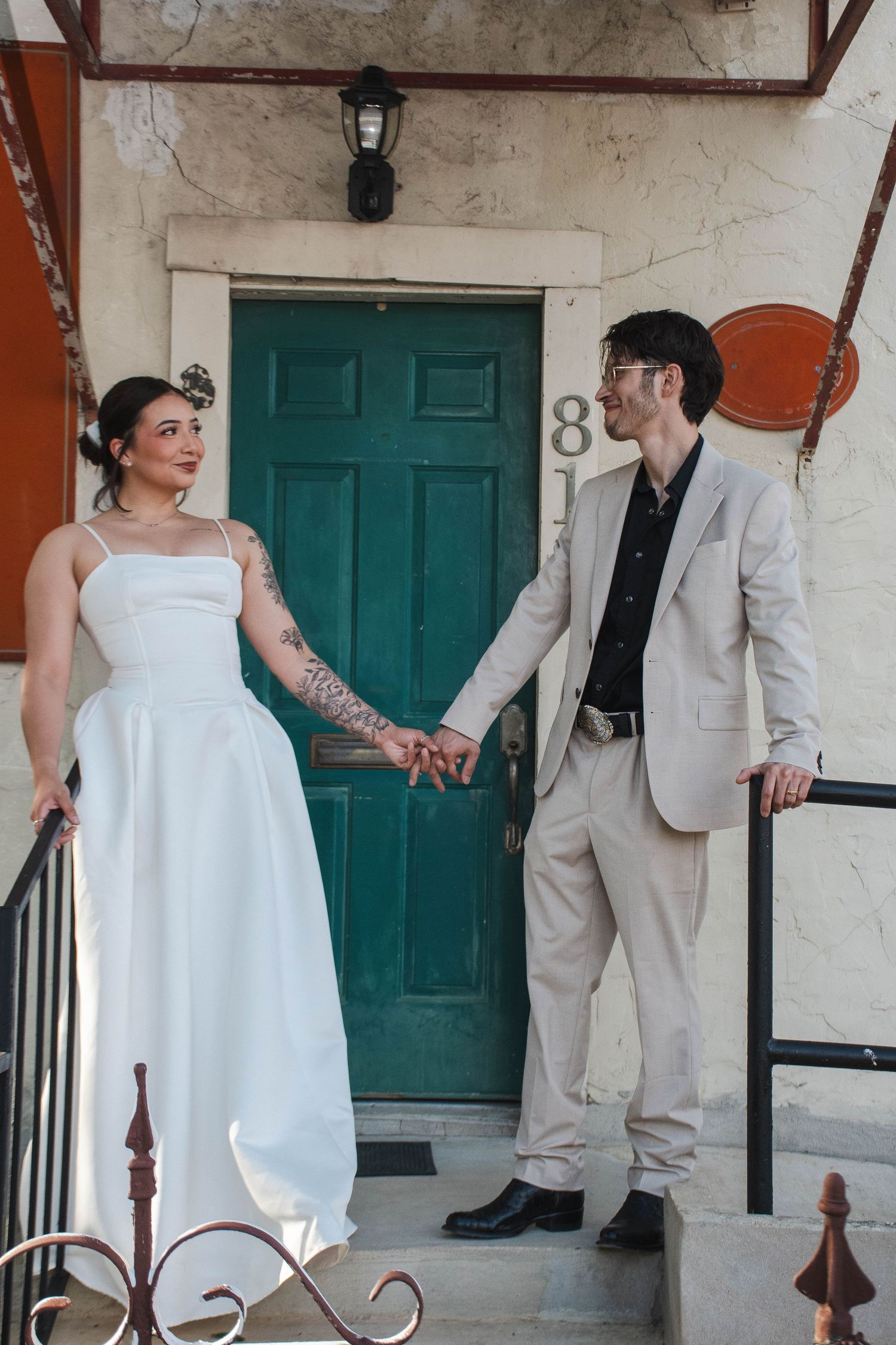 Bride with tattooed arm in white gown and groom in beige suit hold hands before a teal door at number 81.