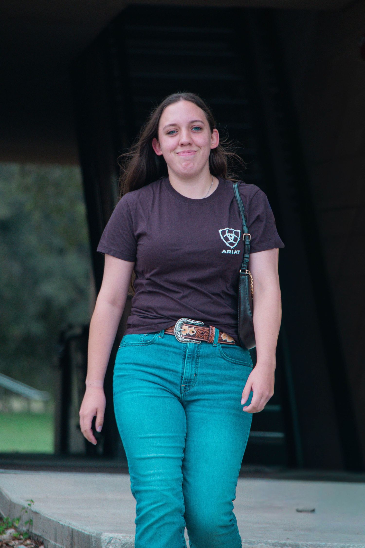 Young woman in teal jeans and dark Ariat t-shirt walking outdoors, smiling with brown hair blowing in wind.