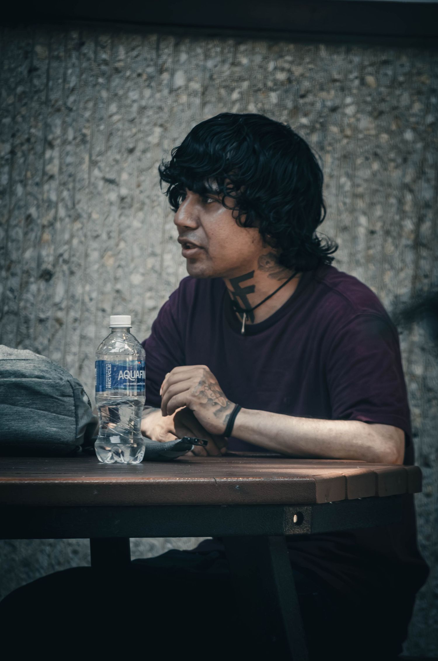 Young man with neck tattoos and curly hair sitting at an outdoor table with a water bottle.