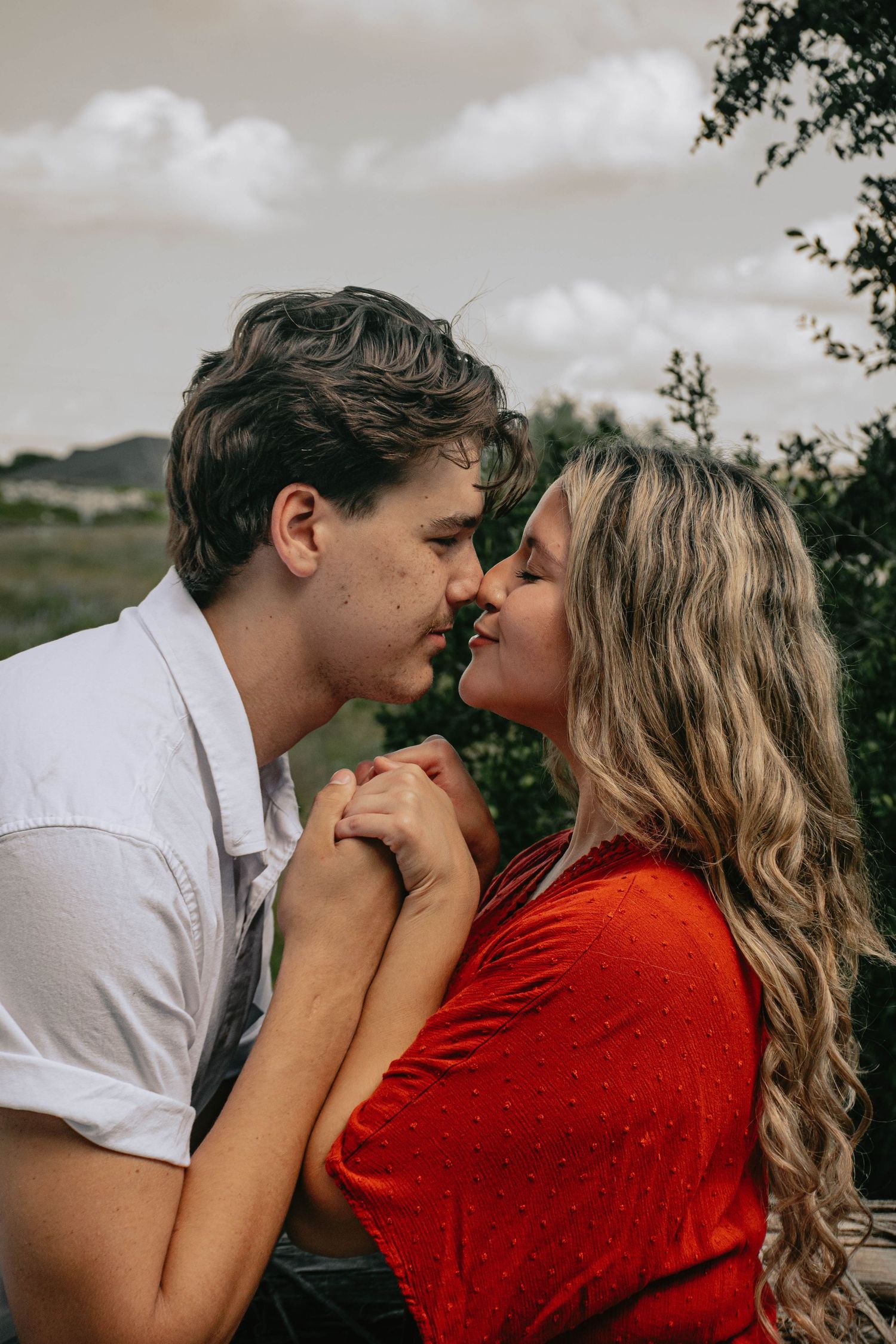 Romantic couple holding hands and sharing a kiss outdoors with greenery in the background.