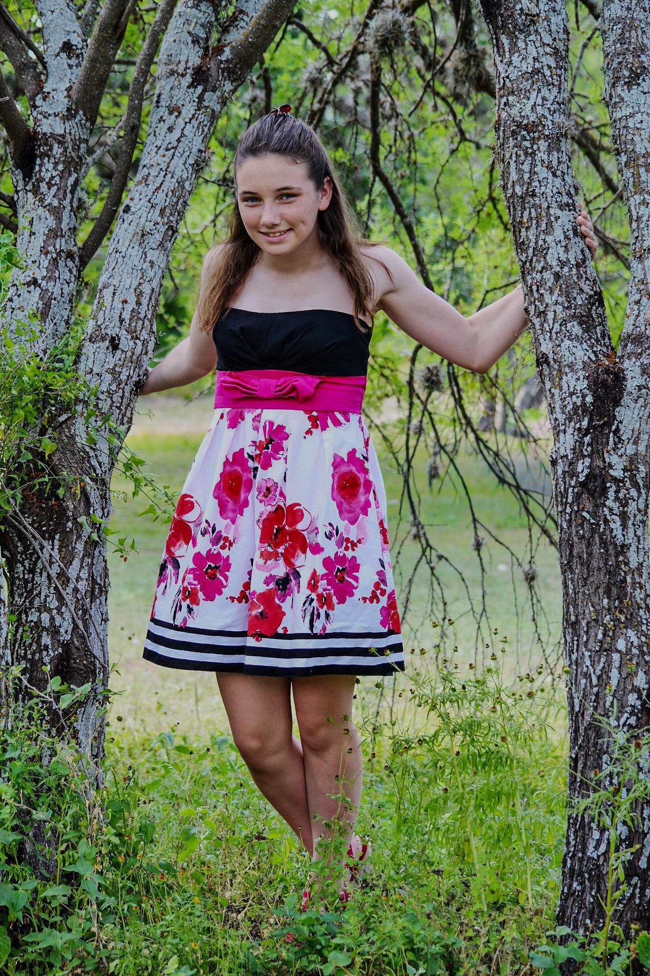Smiling teenage girl in a floral pink and black strapless dress poses between two trees outdoors.