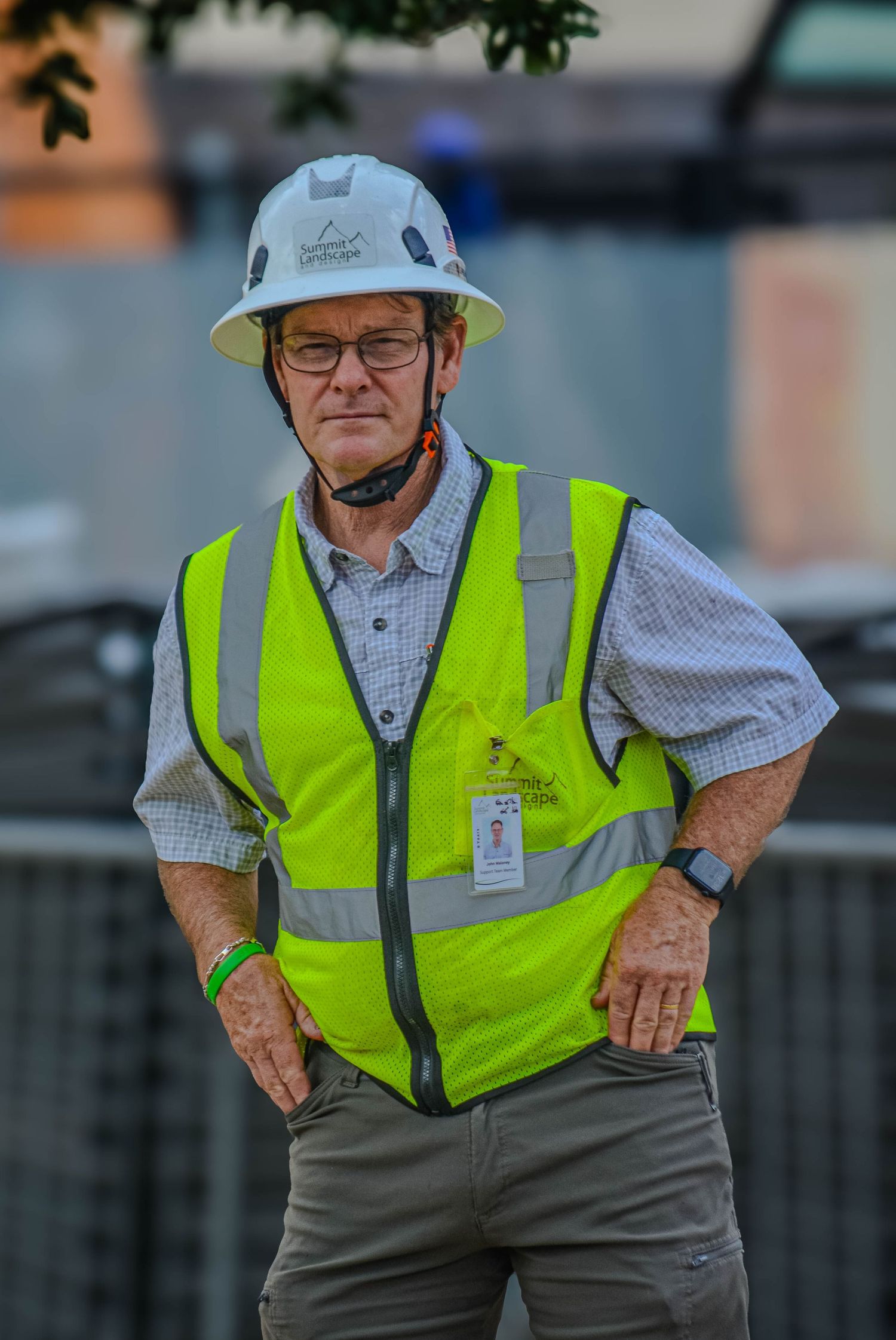 Construction site supervisor wearing yellow safety vest and white hard hat standing with hands on hips.