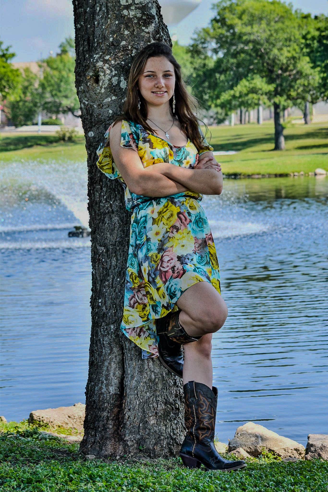 A young woman in a floral dress and cowboy boots leaning against a tree near a pond.