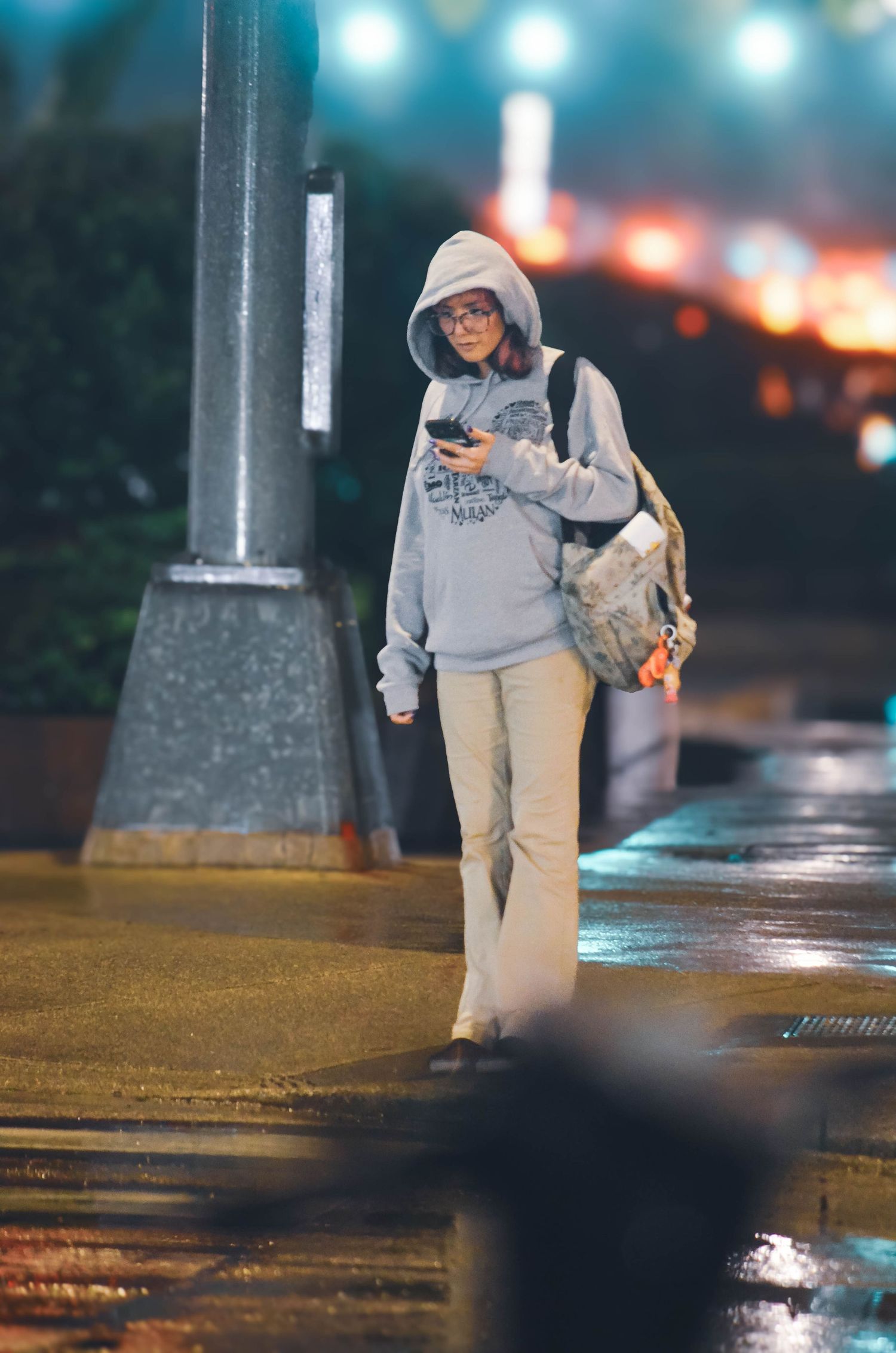 Young woman in a grey hoodie walks at night in the rain while checking her smartphone.