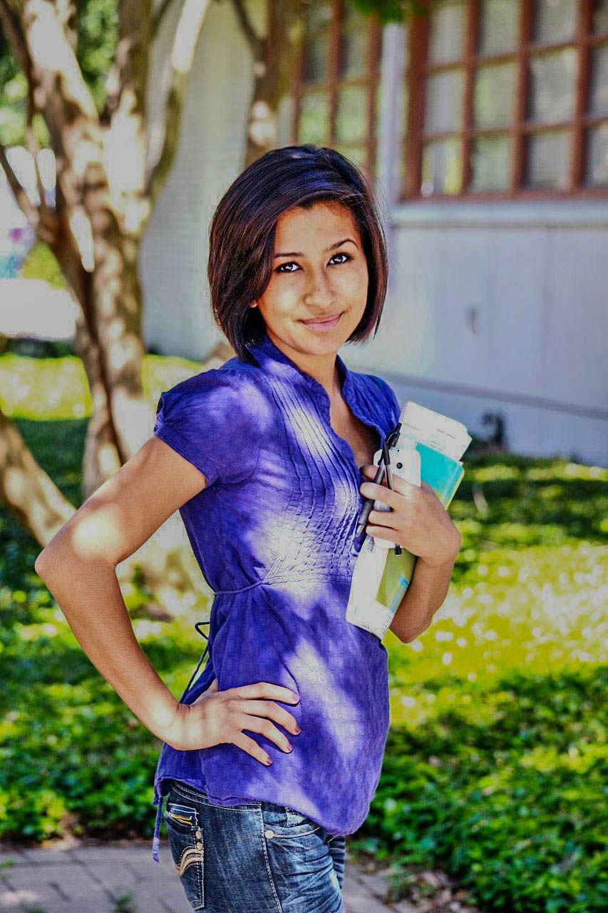 Young woman in purple top holding books smiling outdoors near a building.