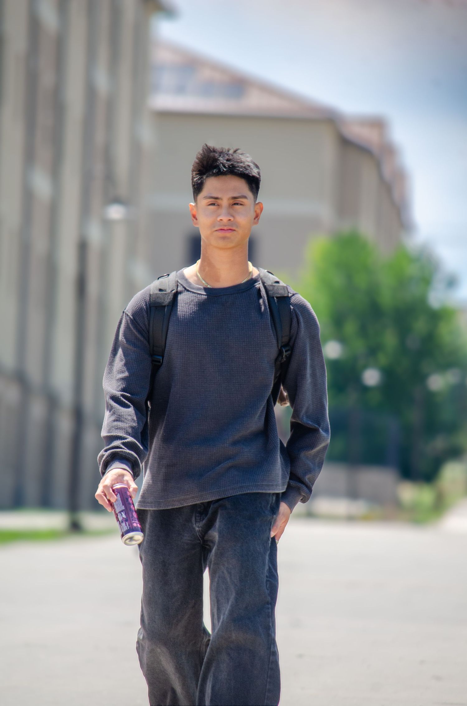 Young Asian male student walking outdoors holding drink wearing navy sweatshirt and backpack.