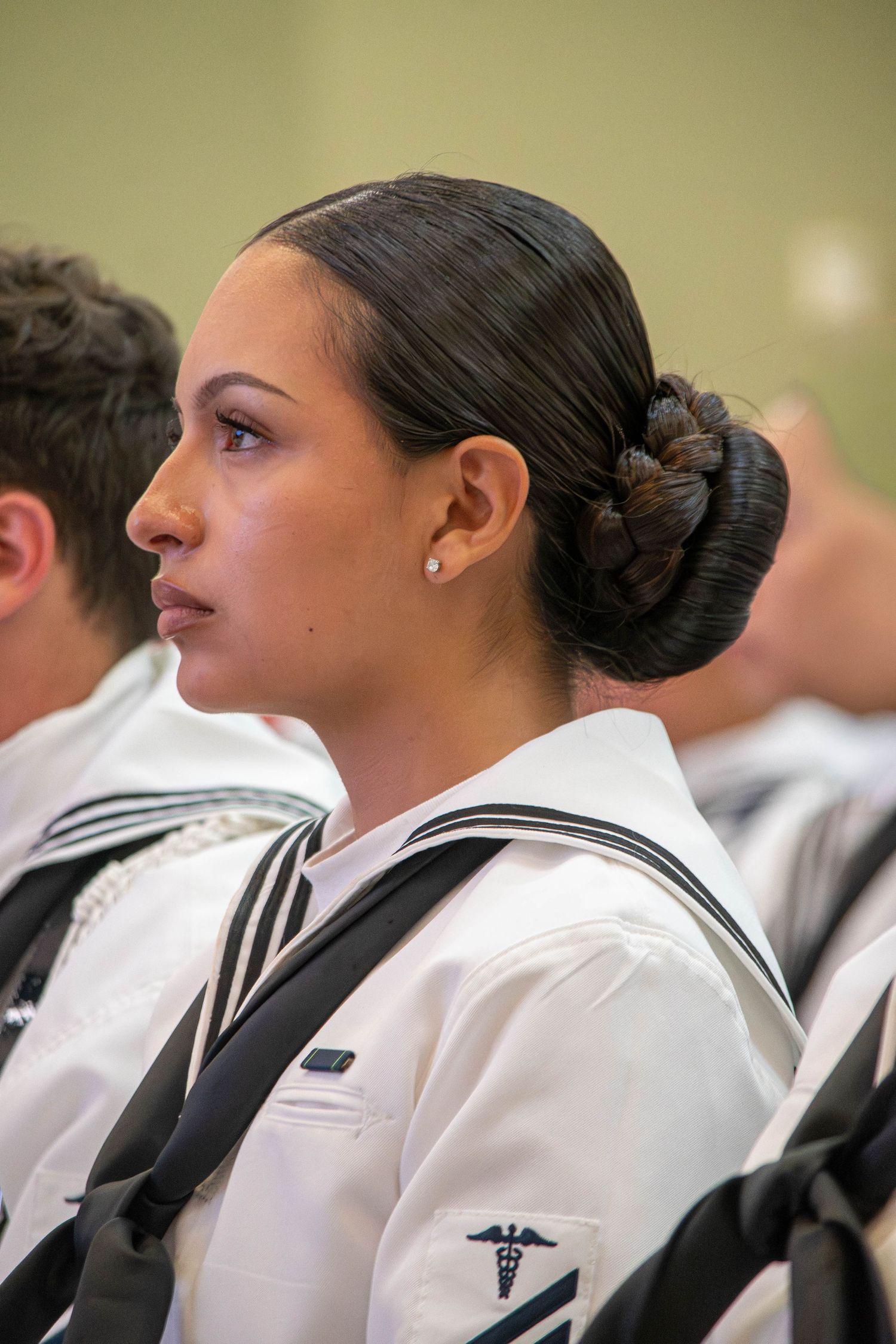 Female Navy sailor in white uniform with medical insignia and bun hairstyle looking attentively forward.