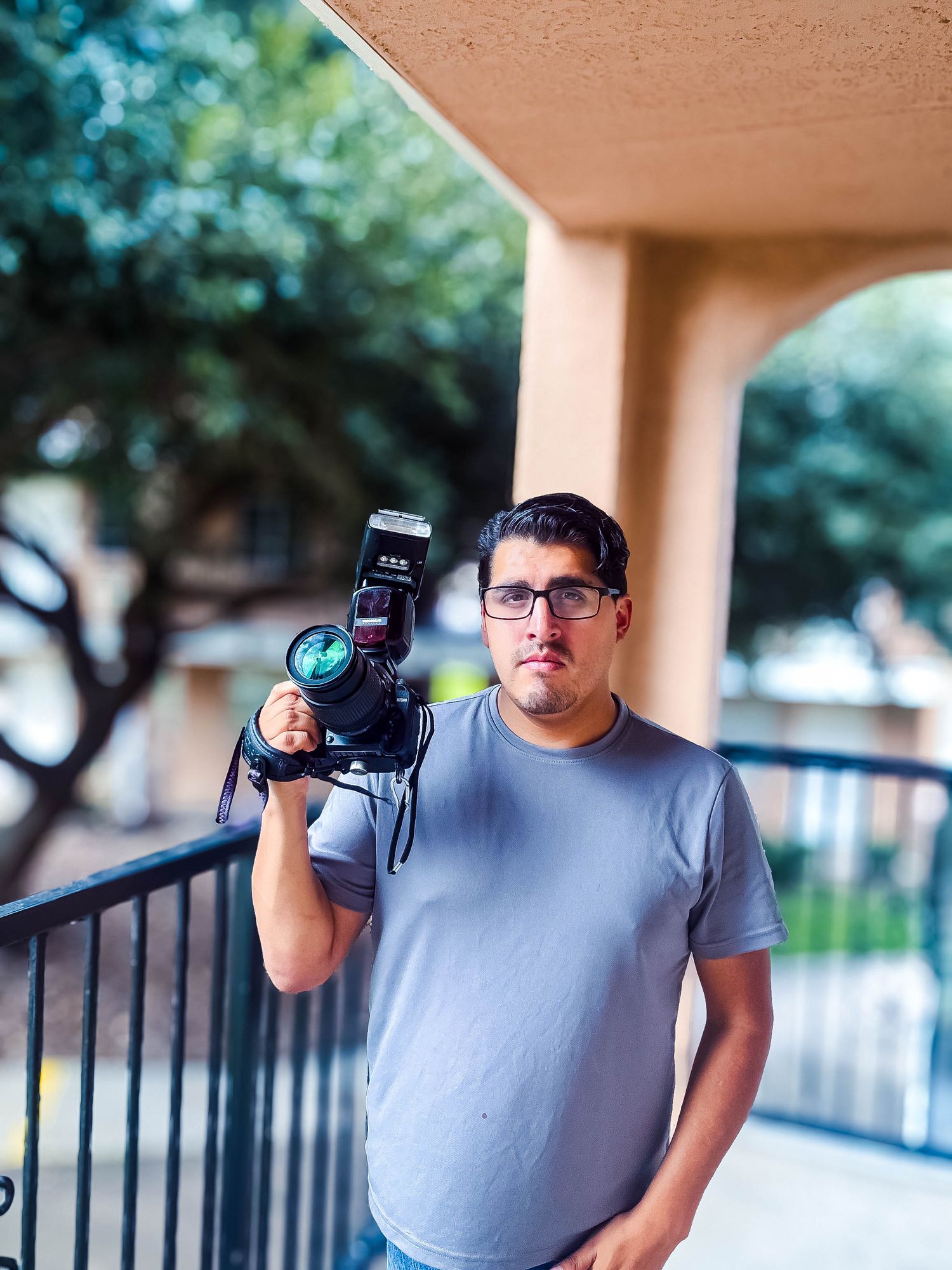 Young man holding DSLR camera with flash on balcony with trees in background.