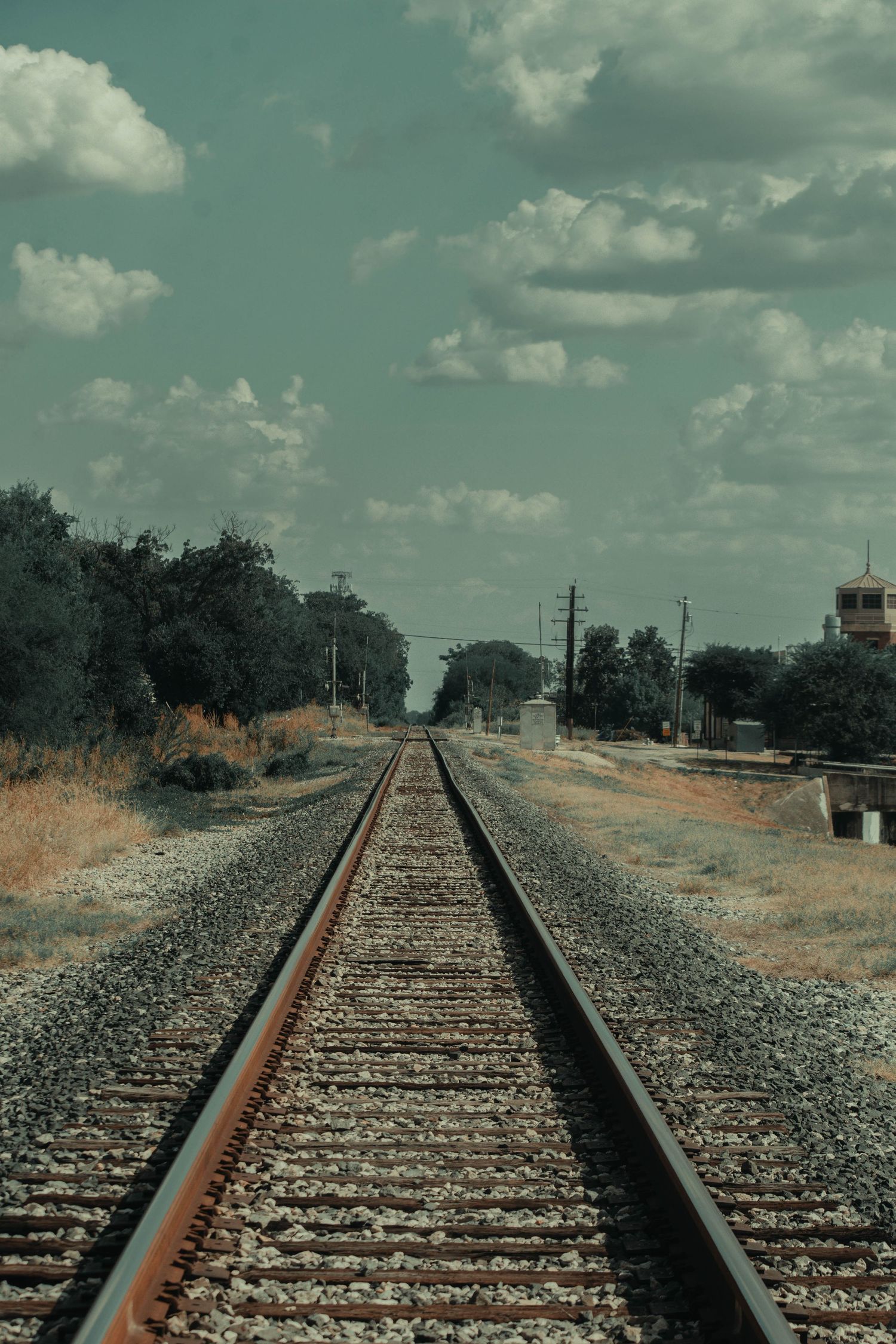 Railroad tracks stretching into the distance through a rural landscape under a partly cloudy sky.
