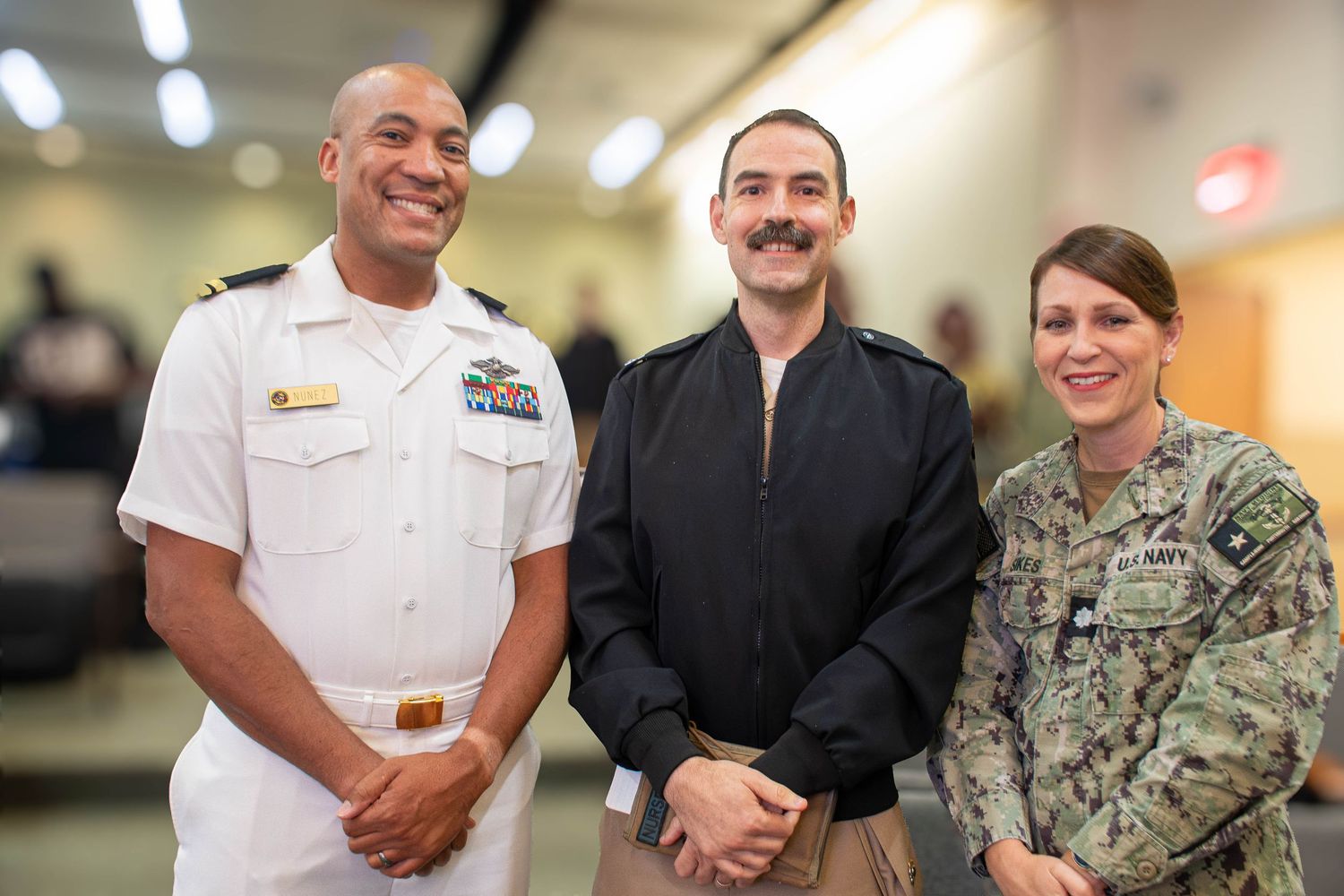 Three smiling military personnel in Navy uniforms pose together at an indoor event.
