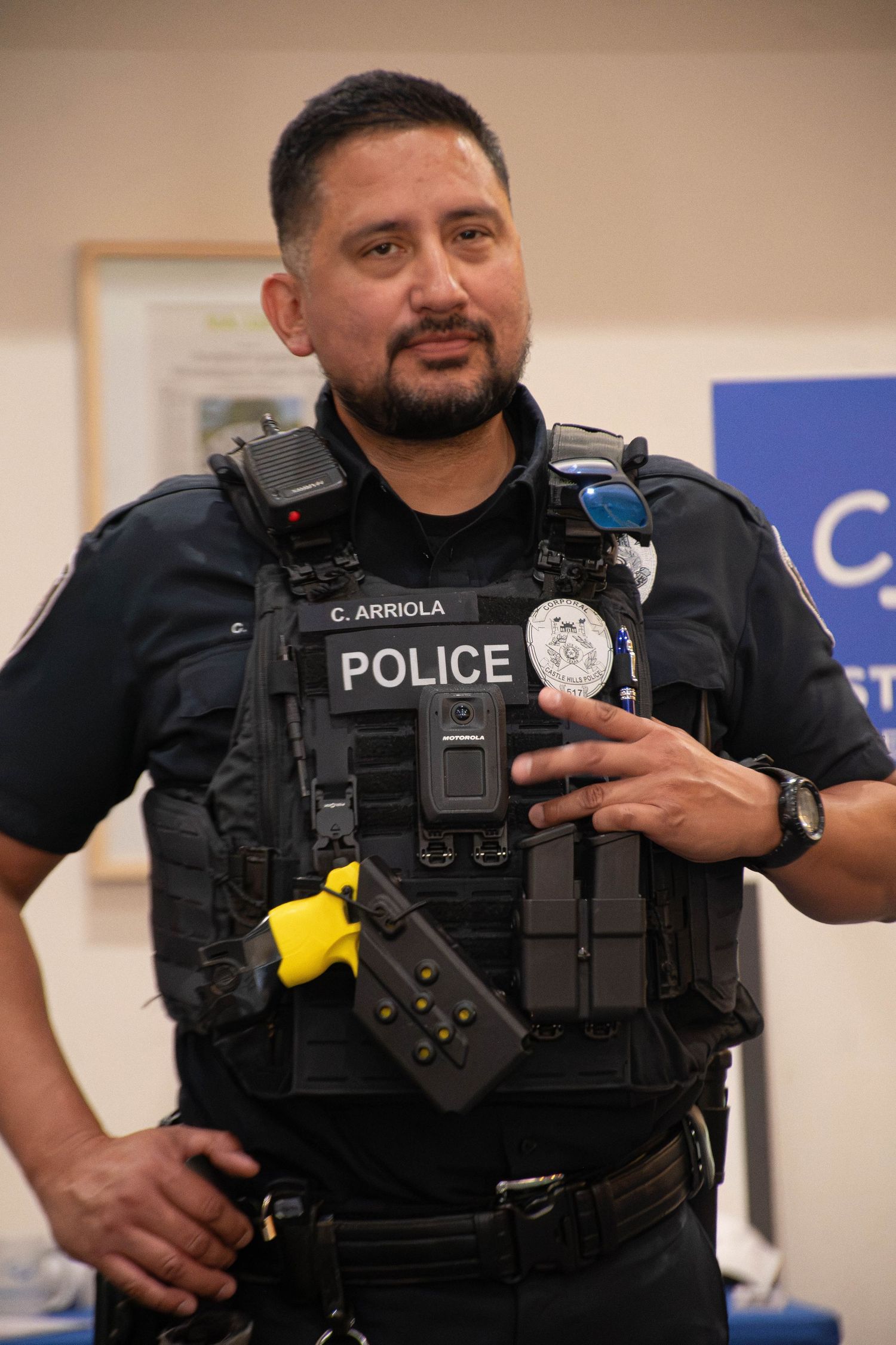 Police officer C. Arriola in tactical vest and gear gesturing indoors.