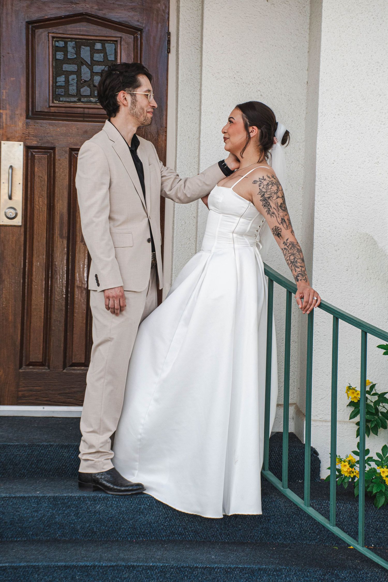 Bride with tattooed arm in white gown and groom in beige suit share a moment on church steps together.