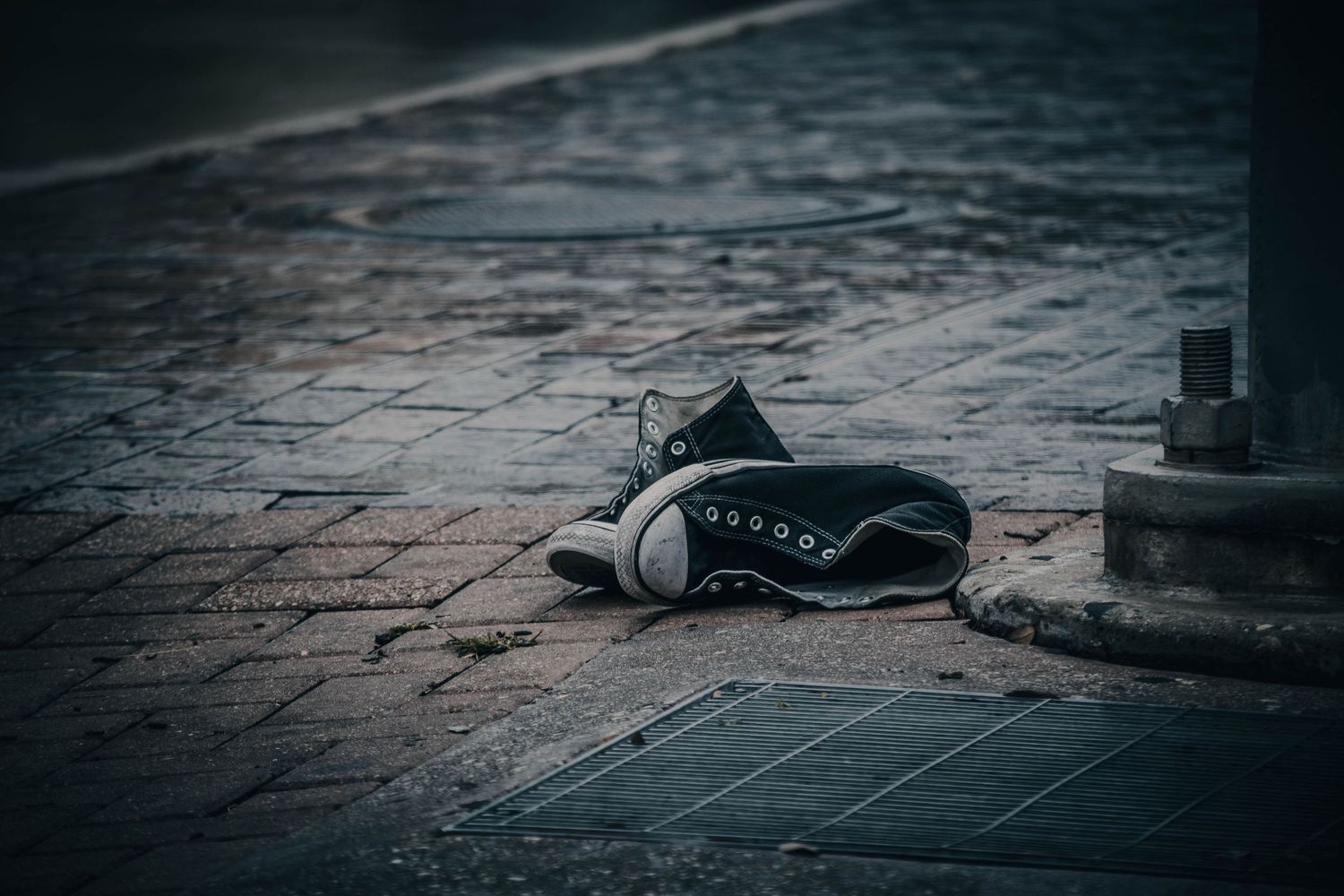 Abandoned black high-top sneakers left on a wet cobblestone sidewalk near a curb.
