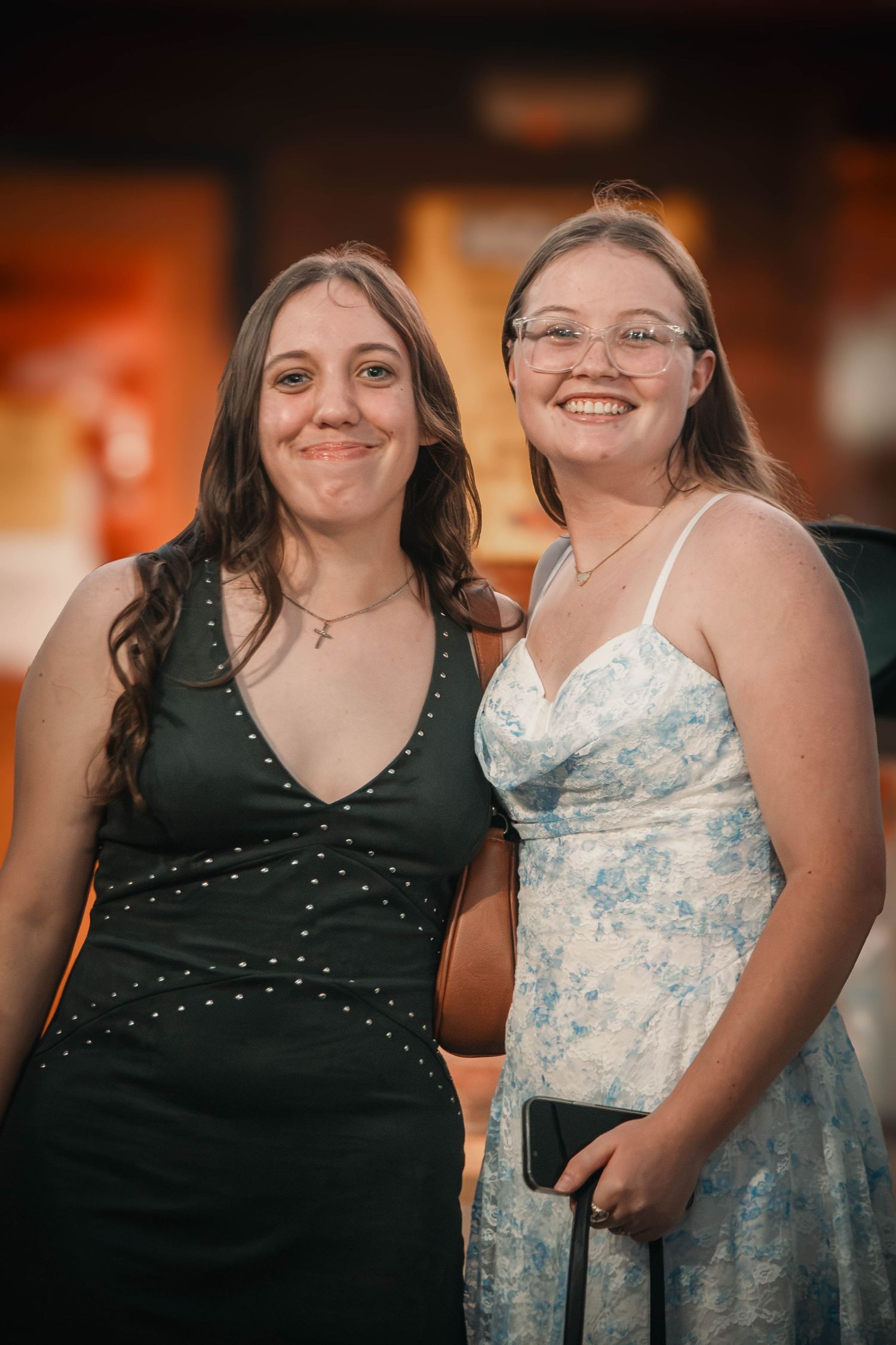 Two young women in formal dresses smiling together, one in black and one in floral blue and white.
