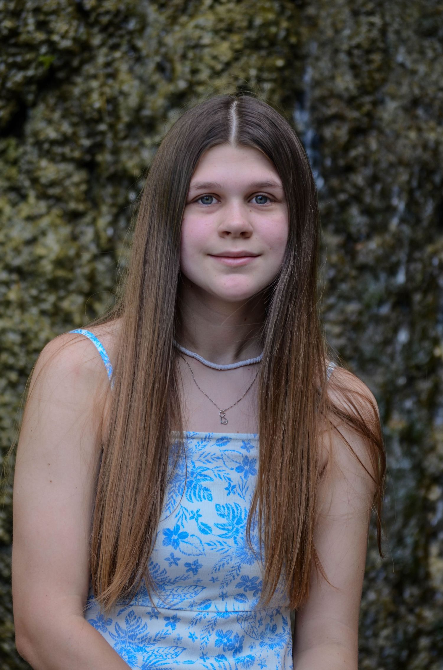 A teenage girl with long straight brown hair wearing a blue floral sundress standing outdoors.